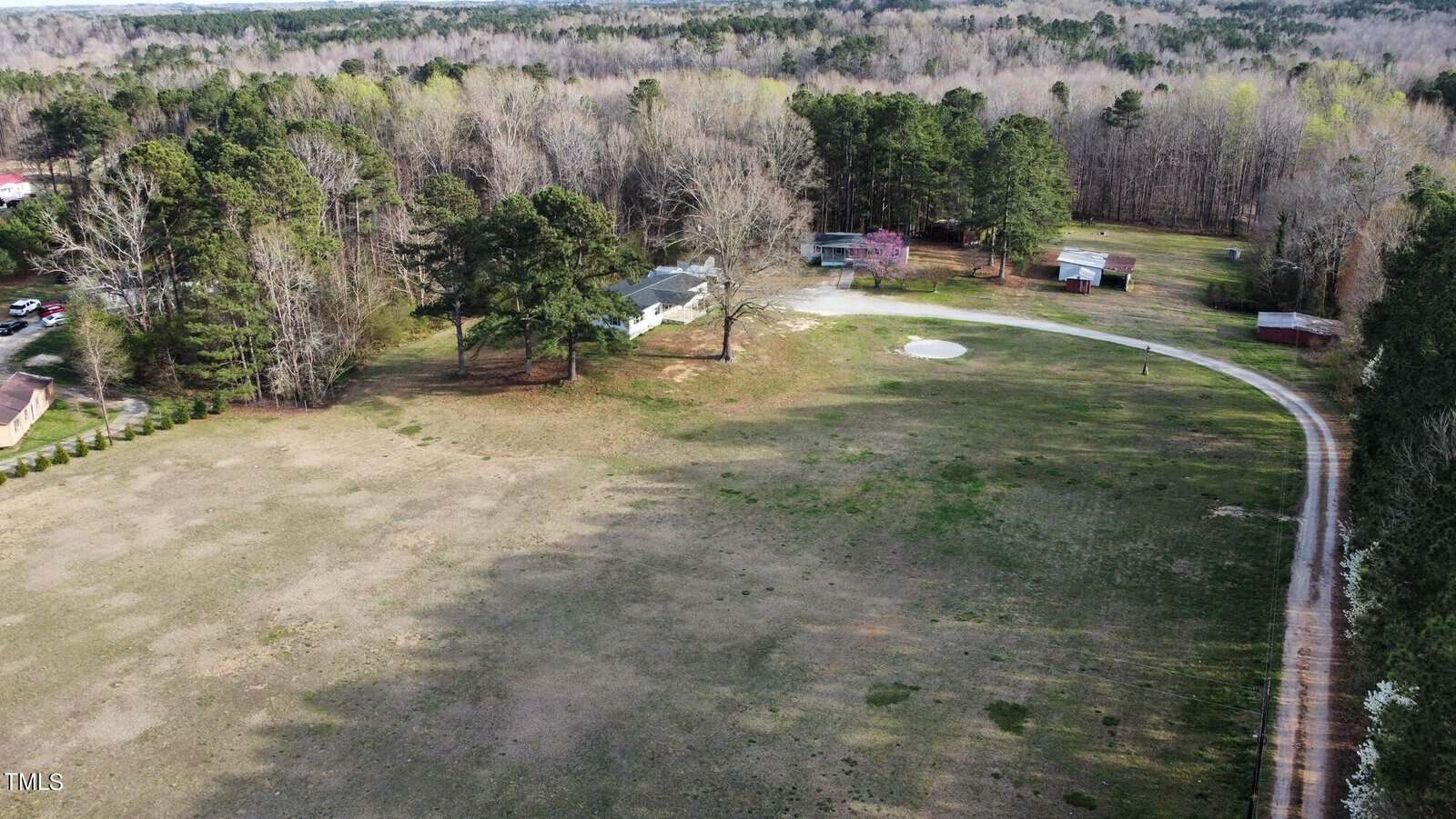 Aerial view of a rural landscape featuring a house surrounded by sparse trees, a large open field, and a curved driveway, with neighboring properties visible in the distance.