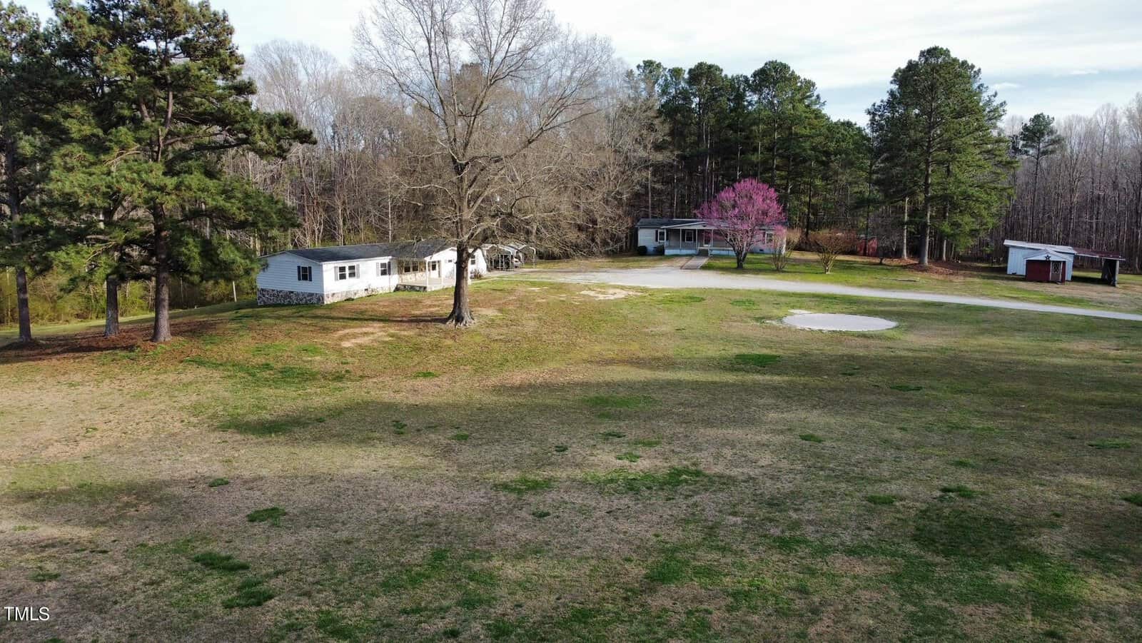 Aerial view of a rural landscape featuring two houses and outbuildings surrounded by scattered trees and a large grassy area. a single pink-flowering tree adds a splash of color.