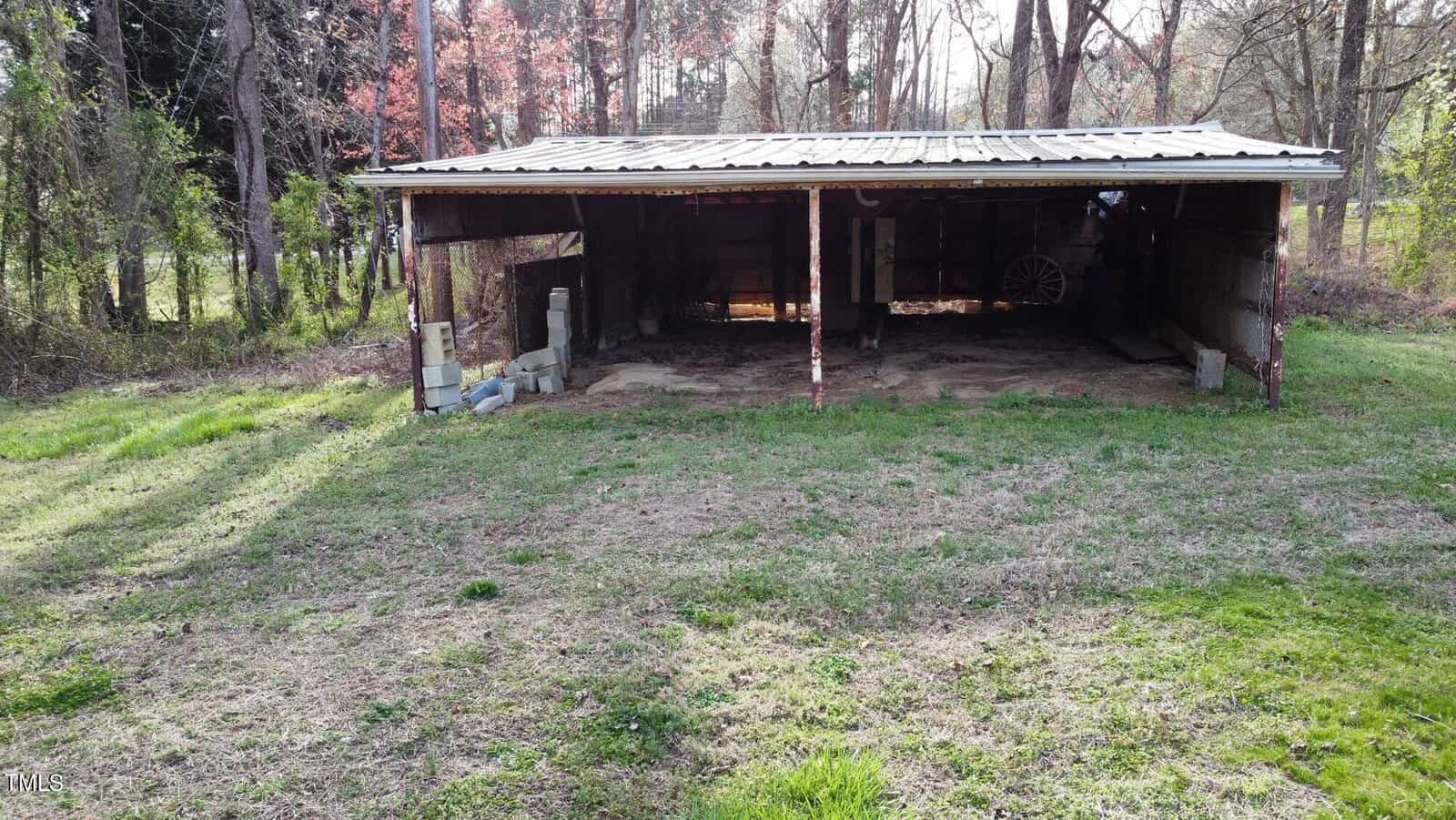 An old wooden shed with a corrugated metal roof, surrounded by grass and trees, partly shaded, housing an antique wagon on the left side.
