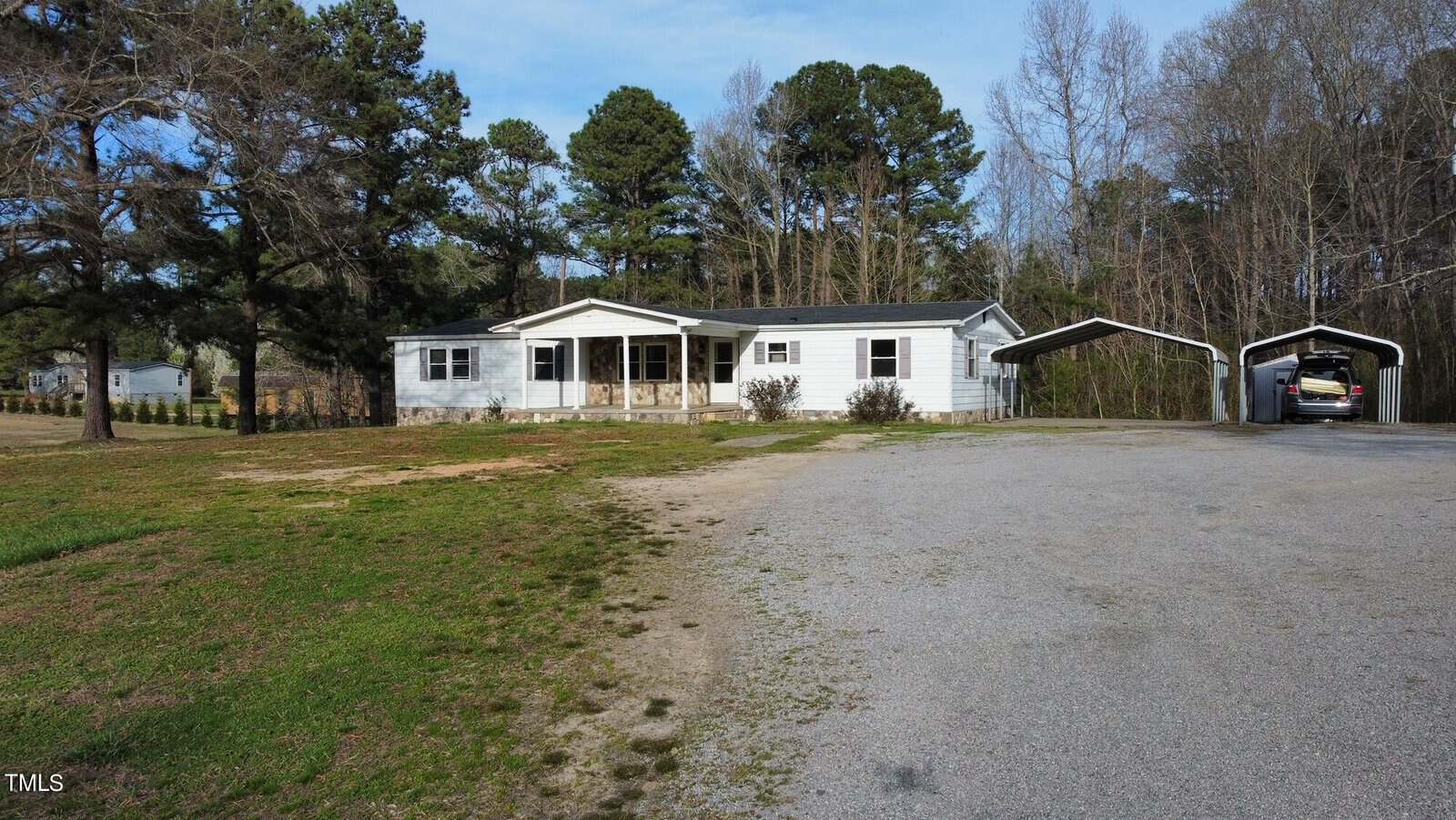 Aerial view of a white single-story house with a covered porch and two-car garage, surrounded by trees and a large grassy area with a gravel driveway.