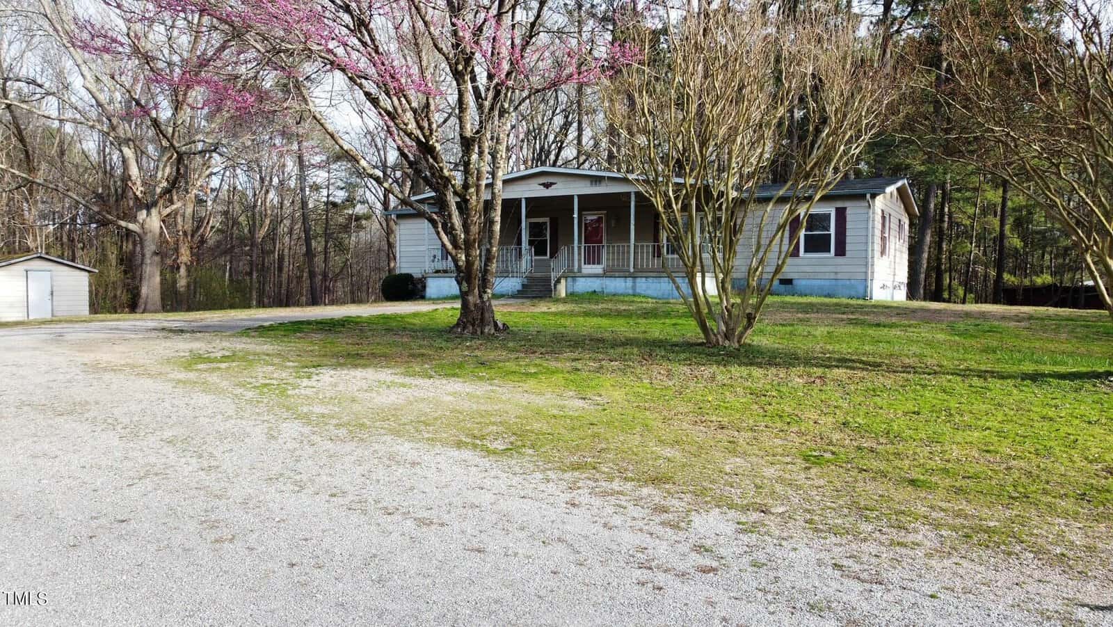 A modest single-story house with a front porch and white siding, surrounded by trees with early spring blossoms. a gravel driveway leads up to the house and a detached garage on the left.