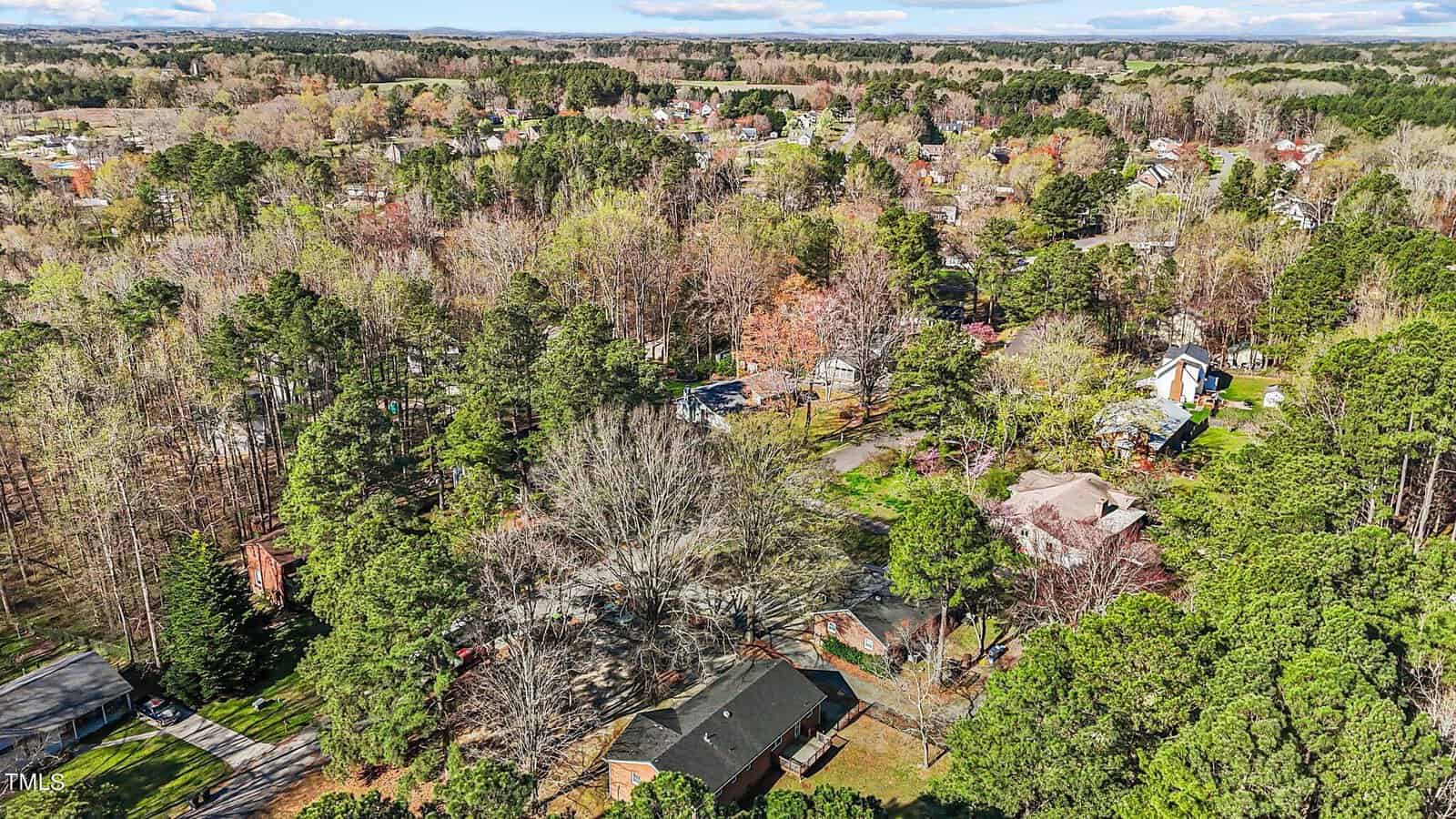 Aerial view of a suburban neighborhood surrounded by dense trees and greenery. Various houses are visible among the foliage, with roads winding through the community. The horizon shows a clear sky and distant landscape.