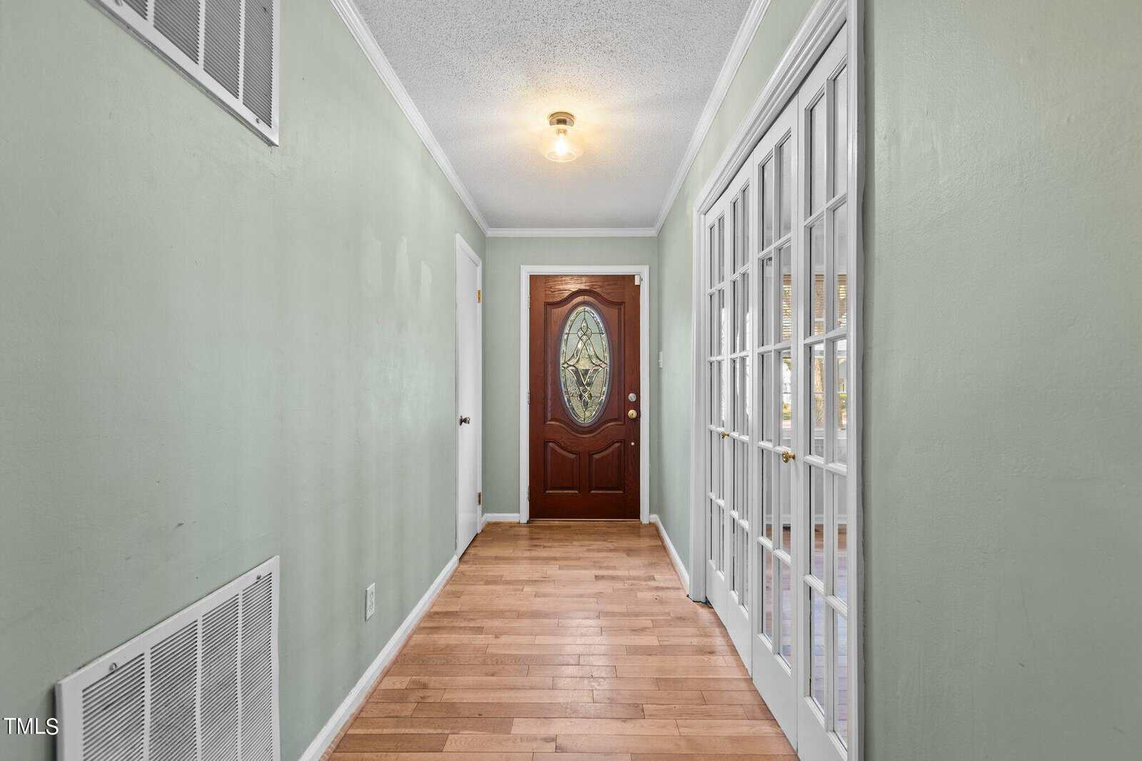 A hallway with light green walls, a wooden floor, and a ceiling light fixture. The hallway features a wooden door with an oval glass design at the end. On the right side, there are white-framed glass panel doors.