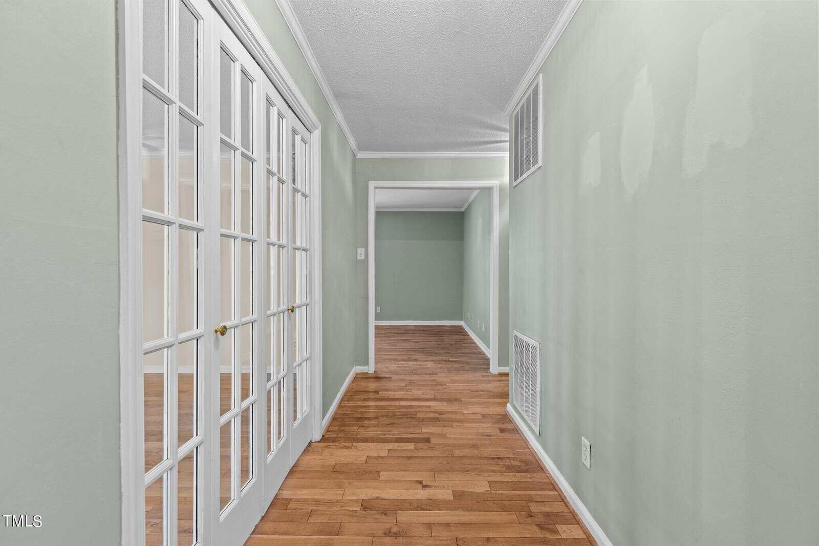 A hallway with light green walls and hardwood floors. French doors open to a room on the left. Two doorways are visible, leading to adjacent rooms, creating a sense of spaciousness.