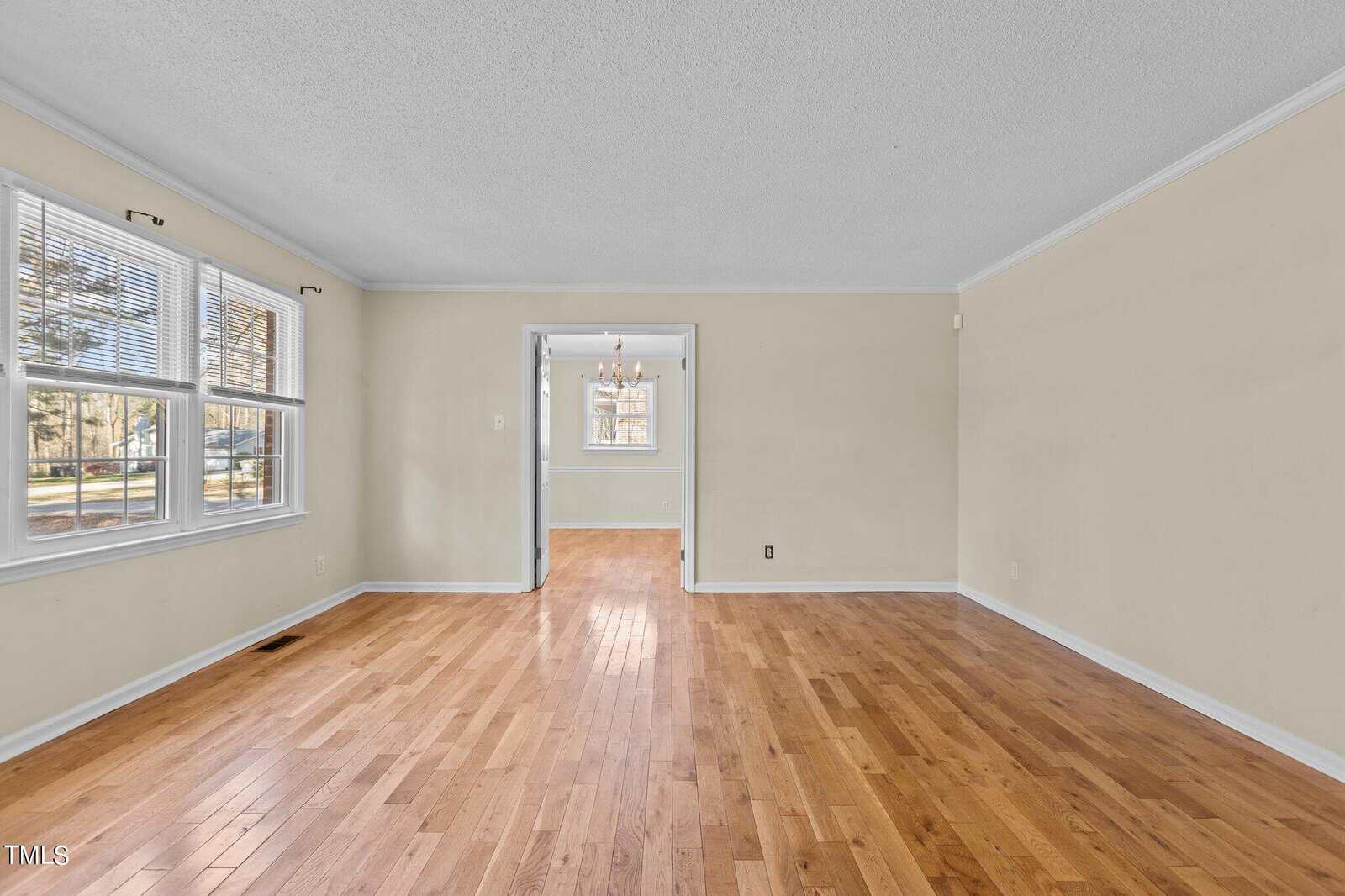 An empty room with light hardwood floors and cream-colored walls, featuring two large windows on the left letting in natural light. A doorway leads to another room with a chandelier.