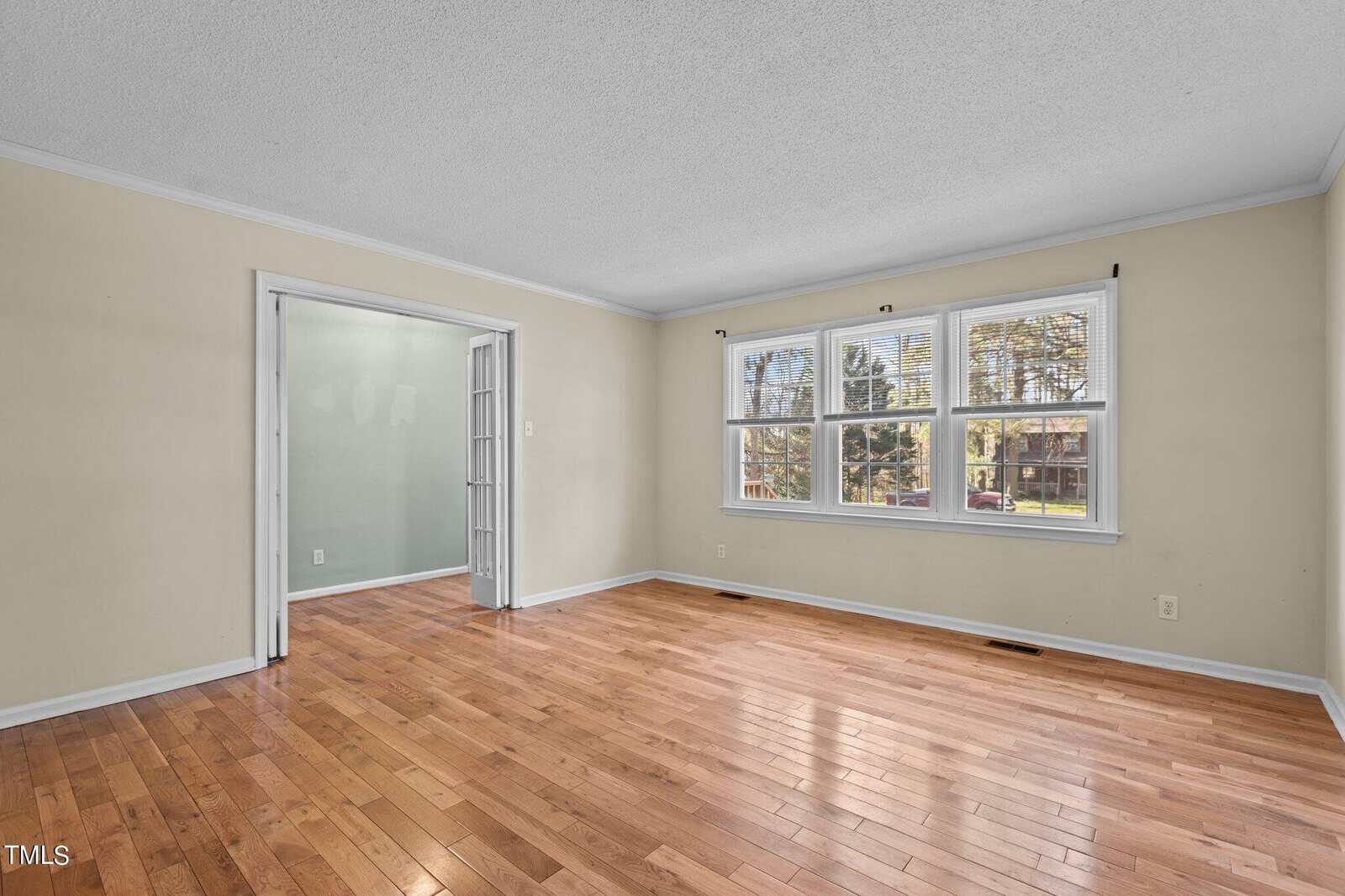 An empty living room with light-colored walls, hardwood floors, and a large window. French doors lead to an adjacent room. Room is well-lit by natural light.