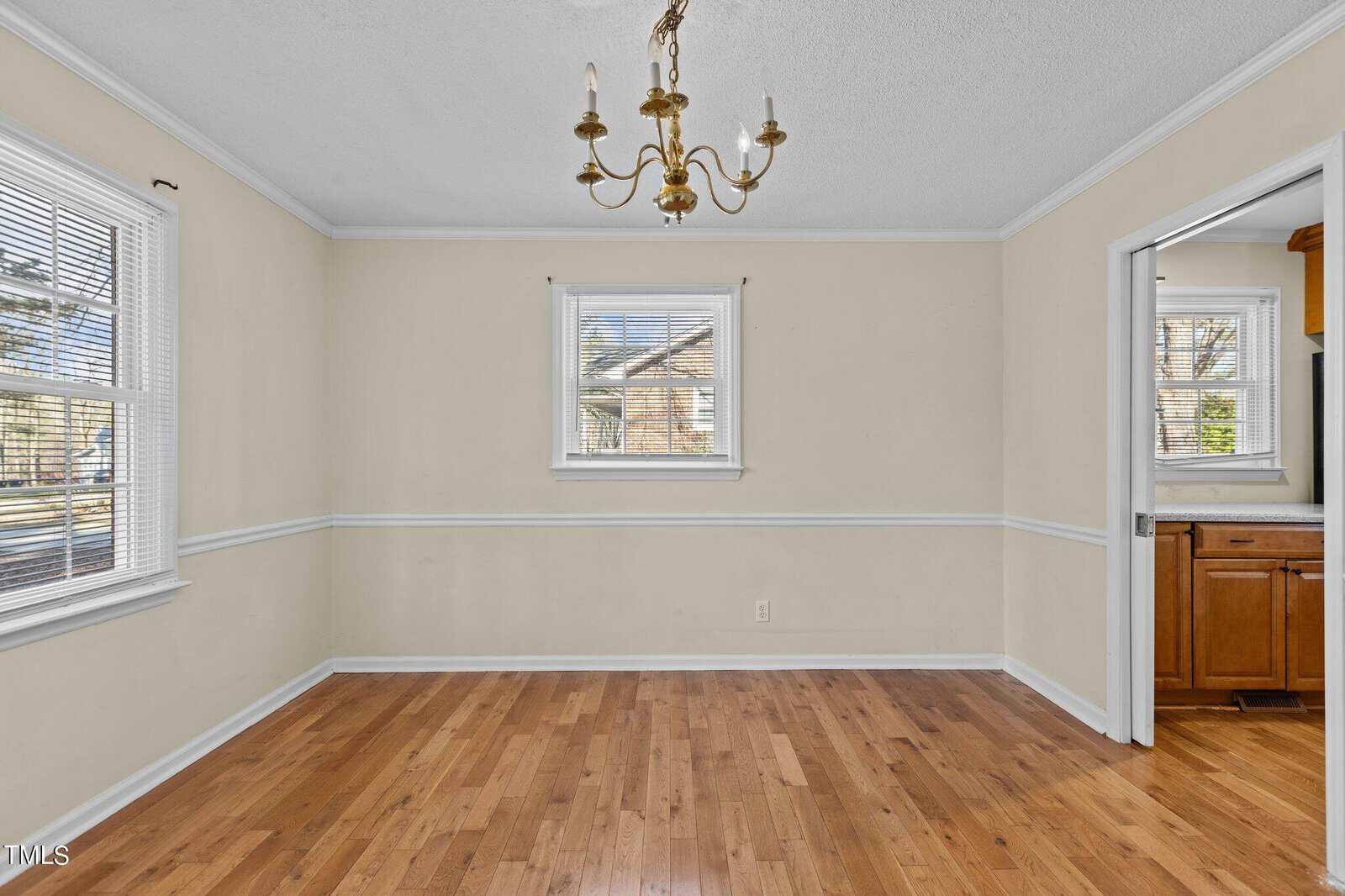 A vacant room with light yellow walls, wood flooring, and a white ceiling. A brass chandelier hangs from the ceiling. There are three windows with white trim, allowing natural light to enter. An open doorway leads to a small kitchen area.