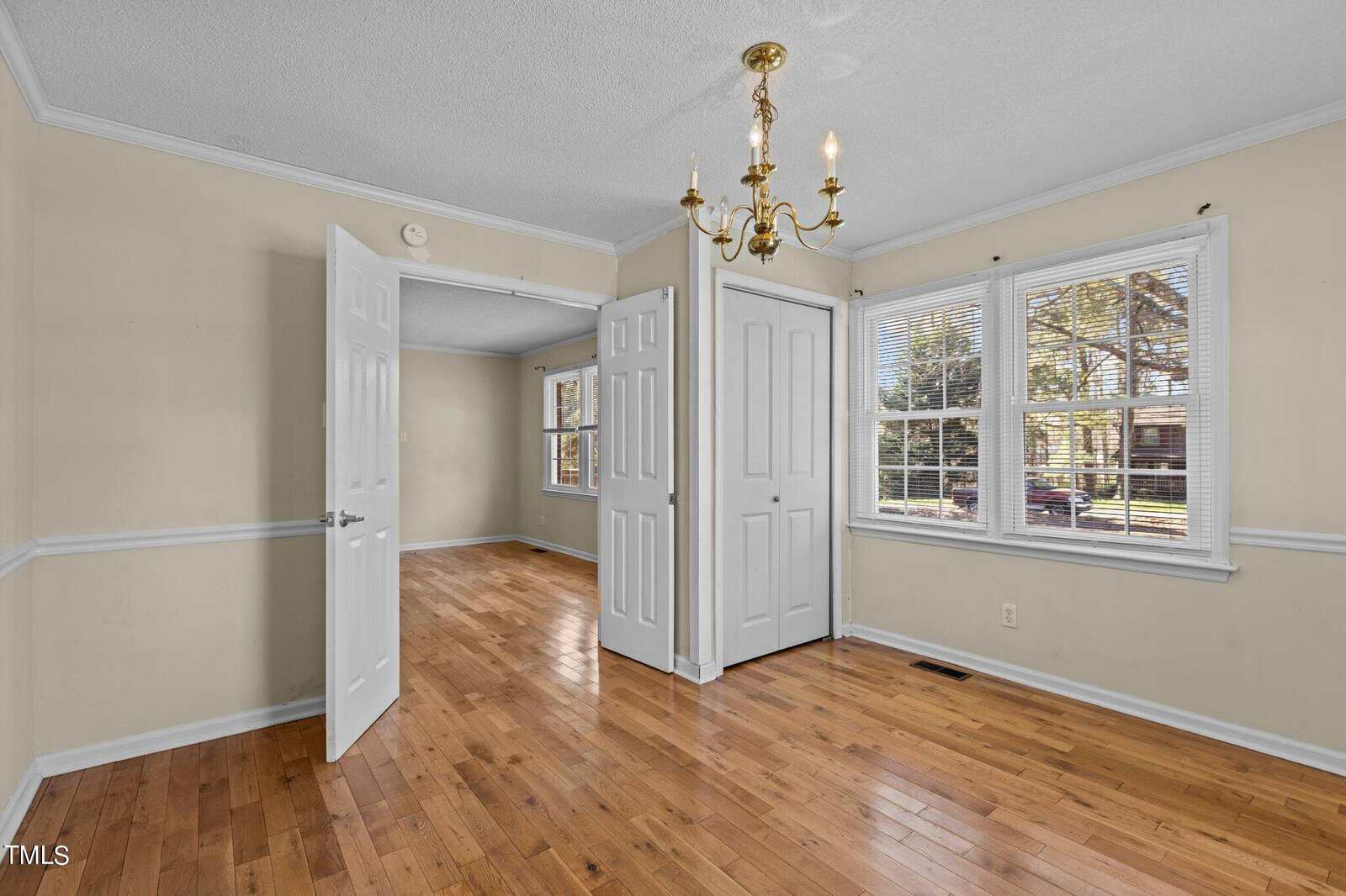 An empty room with light-colored walls and wooden floors. The room features a chandelier, a large window with blinds, double doors, and decorative molding. Sunlight streams in, casting shadows on the floor.