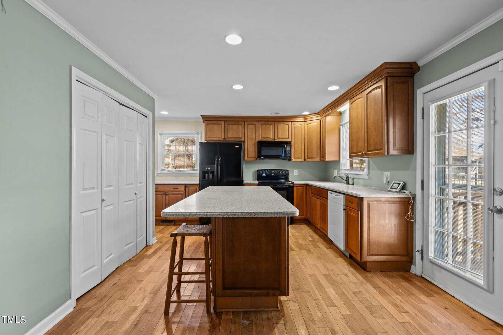 A spacious kitchen features wooden cabinets, a central island with stools, and modern appliances including a black fridge and stove. Light green walls and a door with glass panels enhance the bright and airy feel. Wood flooring adds warmth.