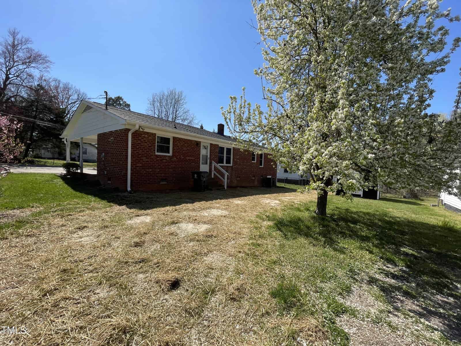 A red brick one-story house with a white carport on a sunny day. the house is surrounded by a grassy yard with a blooming white tree on the left.