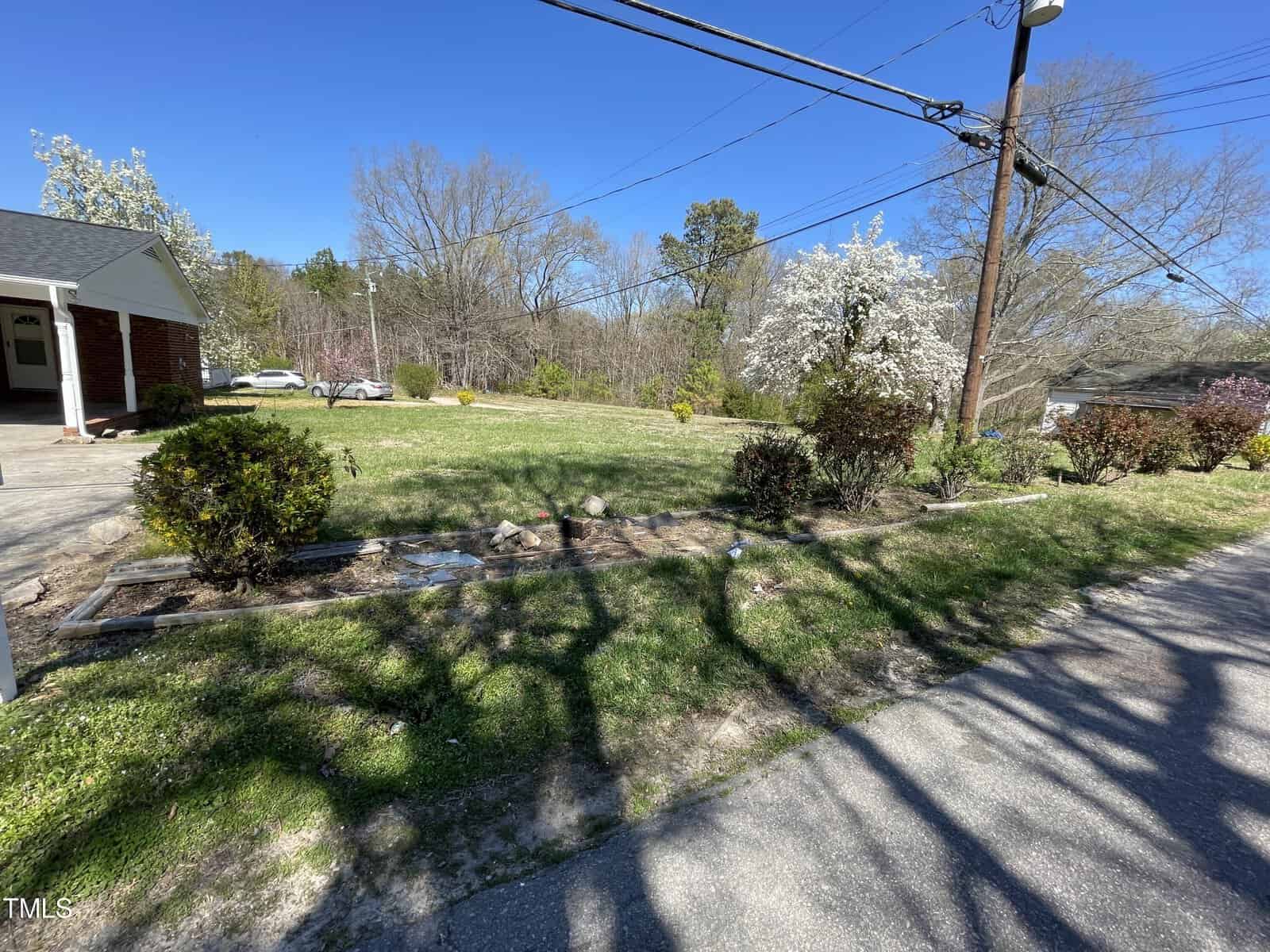 A sunny suburban street scene with a small front yard leading to a single-story house, blooming trees visible, and shadows of utility poles cast on the pavement.