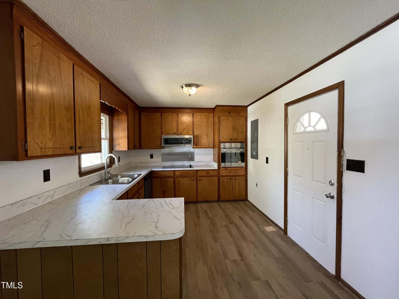An empty kitchen featuring wooden cabinets, white countertops, a built-in microwave and oven, with laminate flooring and a white door on the right.