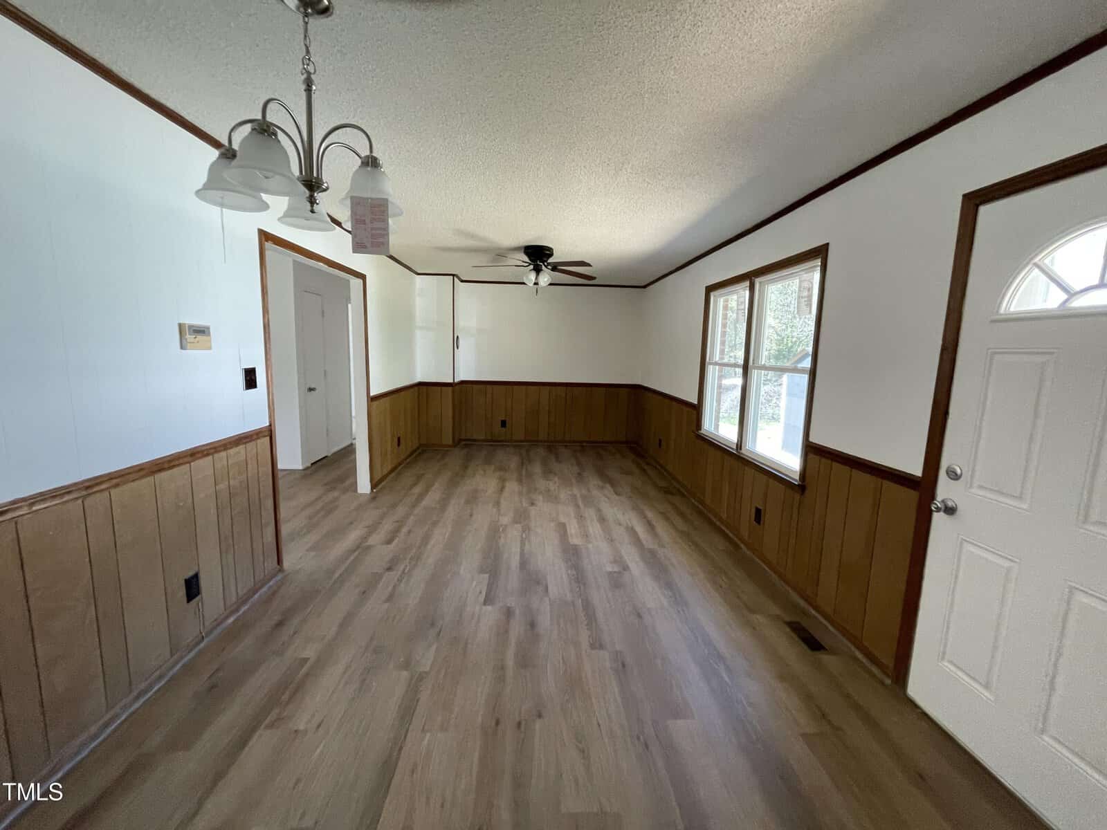 A bright, empty room with wood paneling halfway up the walls, laminate flooring, and multiple windows. includes a ceiling fan and two ornate light fixtures.