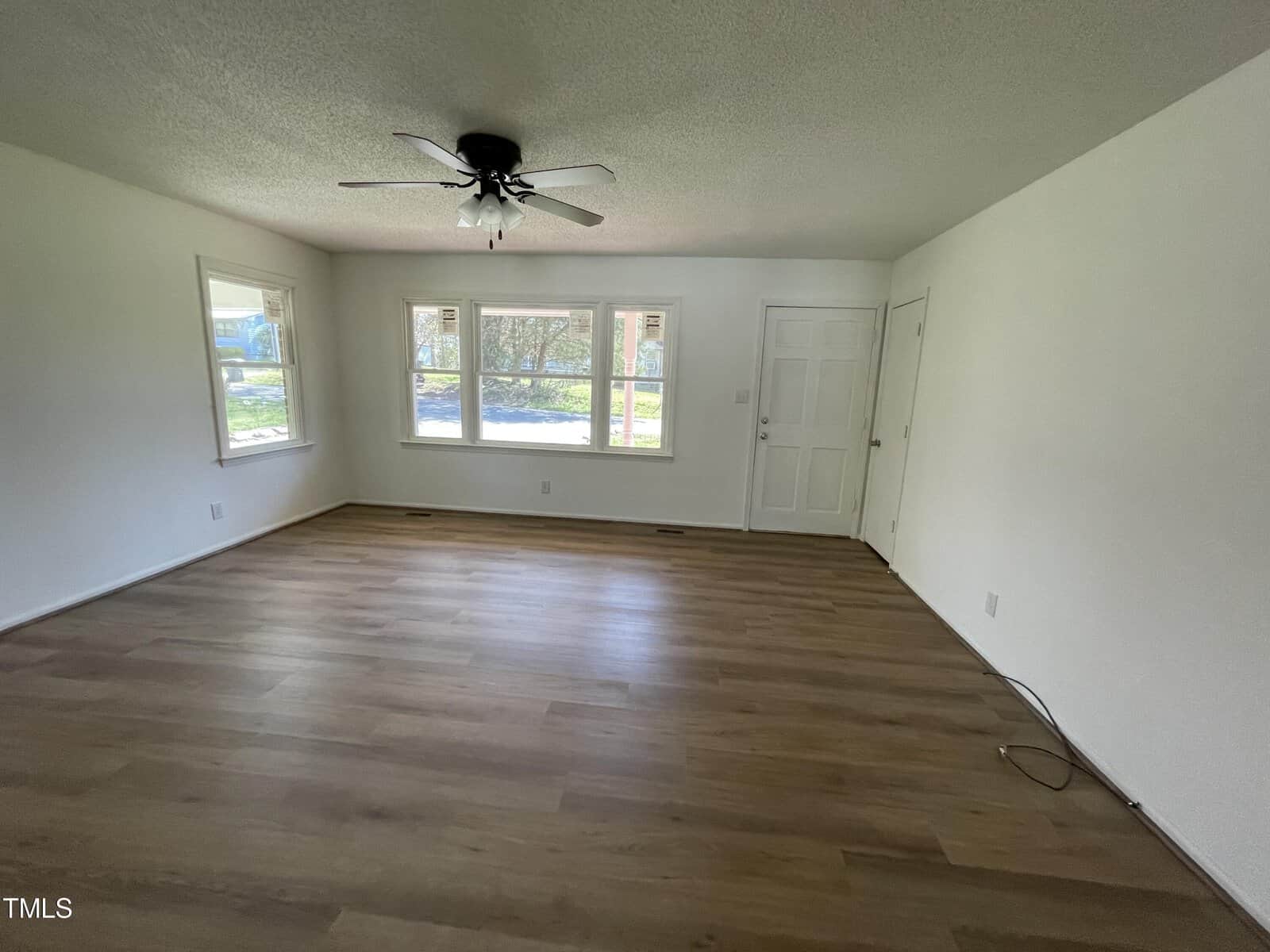 Empty living room with laminate flooring, white walls, ceiling fan, and two windows providing natural light, with a closed white door.