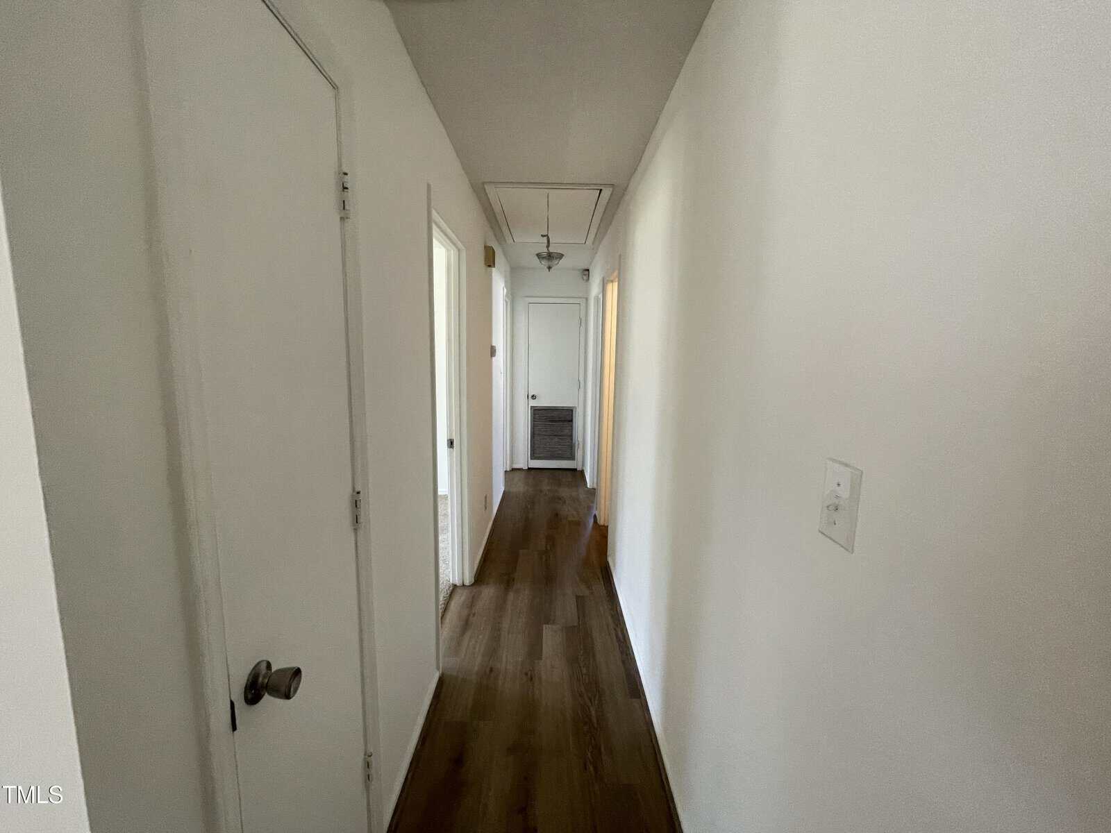 A narrow hallway in a house with white walls and wooden flooring. there are multiple doors on either side, and the hallway ends with a doorway that appears open.