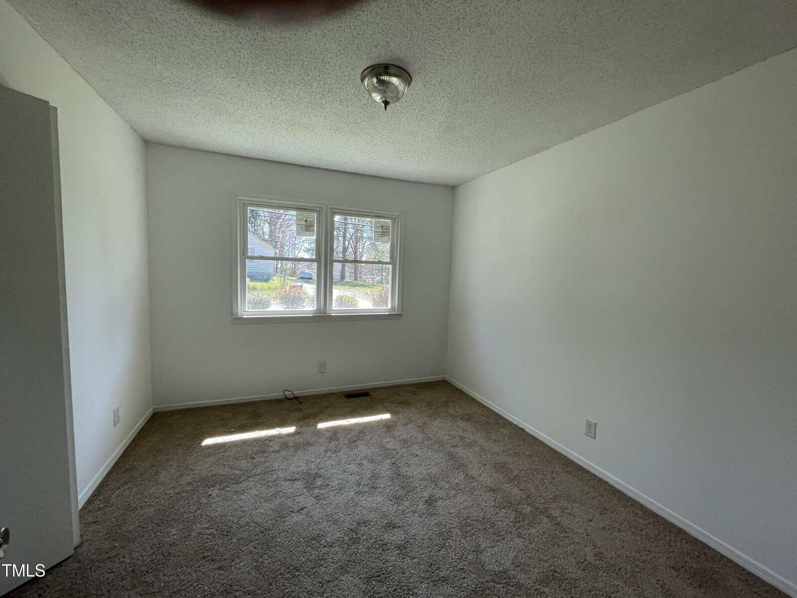 Empty room with beige carpet, white walls, and a double window allowing natural light into the room. a light fixture hangs from the textured ceiling.