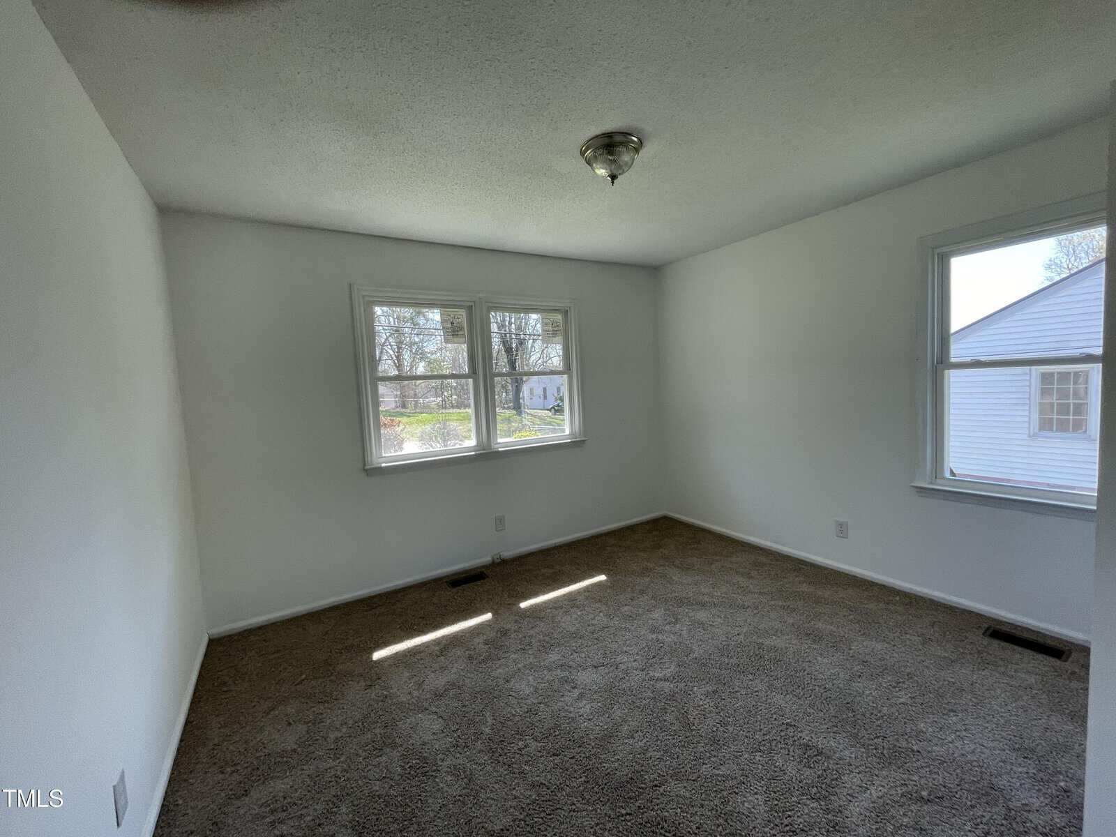 Empty room with white walls and a gray carpet, featuring two windows with a view of neighboring houses. bright natural light illuminates the space.