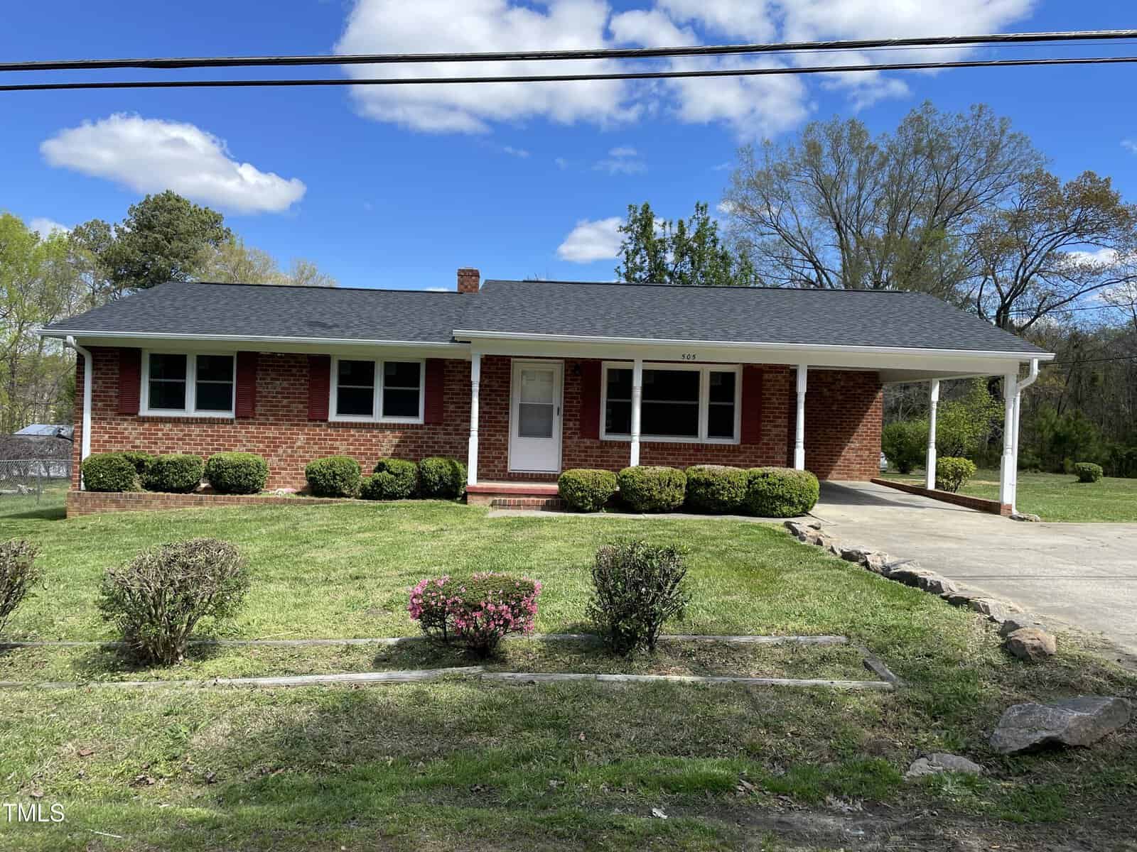 A red brick single-story house at 505 Powell, Henderson NC, with a white porch, surrounded by a well-kept lawn and shrubbery under a clear blue sky.
