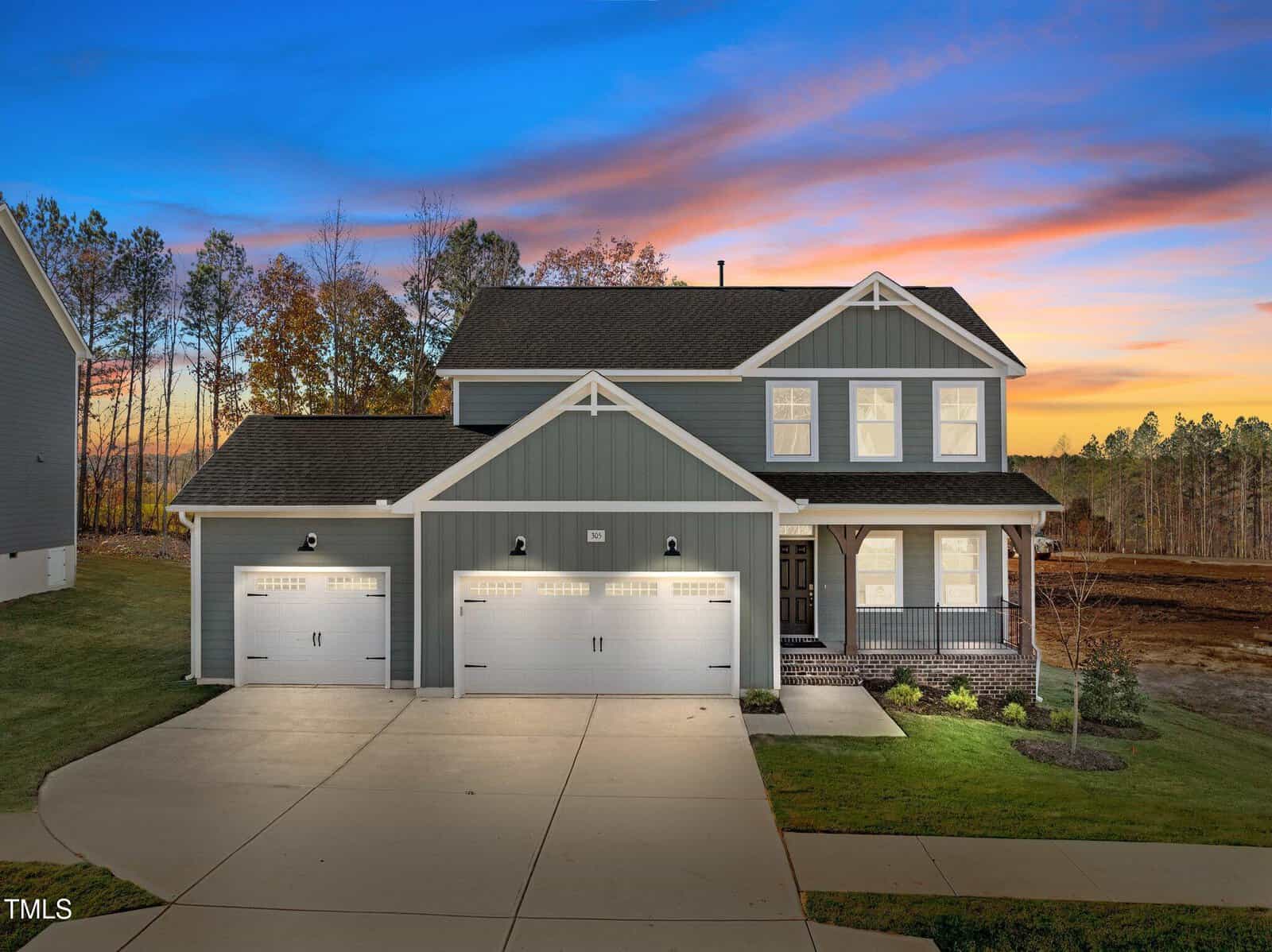 Two-story modern house with gray siding, white trim, and three-car garage, illuminated at dusk. Large driveway, covered front porch, and grassy lawn with a backdrop of trees and a colorful sunset sky.
