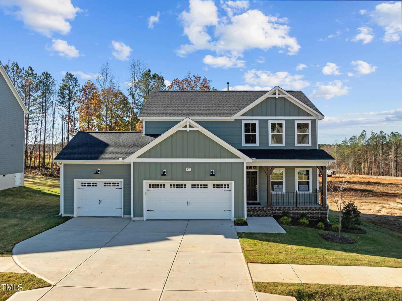 A two-story blue-gray house with white trim, a covered front porch, and three-car garage, sits on a spacious lot with a driveway and lawn, surrounded by trees and open land under a partly cloudy sky.