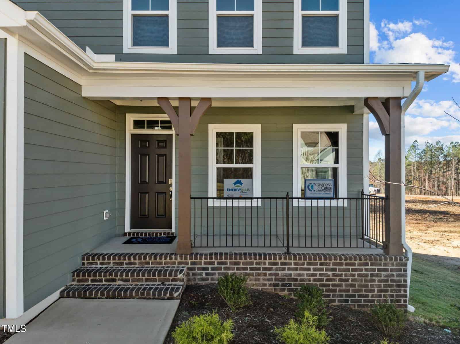 A modern house exterior with green siding, black front door, covered porch with brown wooden posts, brick steps, metal railing, and two windows displaying energy and builder signs. The yard is partially landscaped.