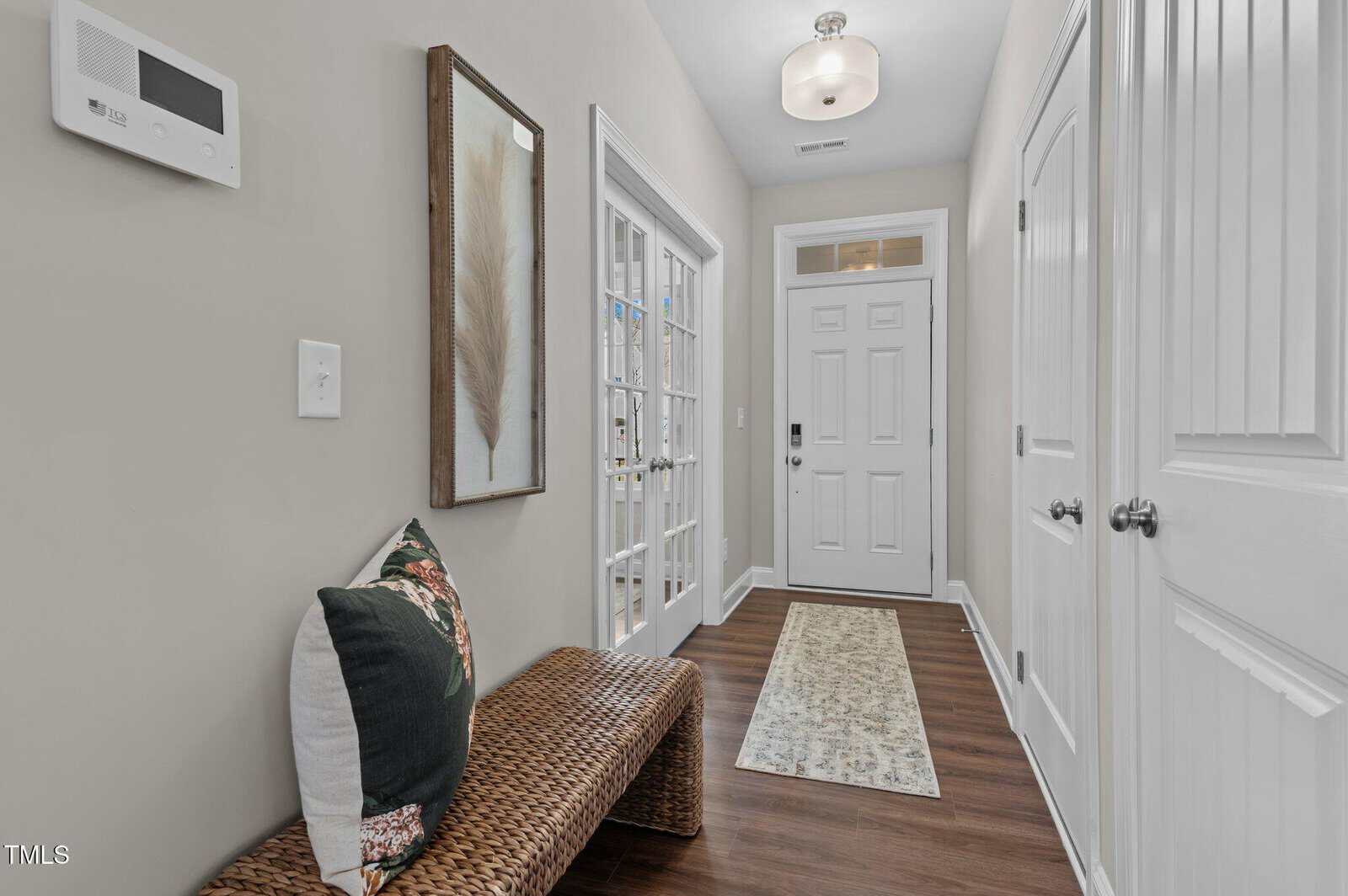 A hallway with white walls and wood floor, featuring a white front door, a woven bench with pillows, a framed feather artwork, a small rug, and two sets of white doors on the right.