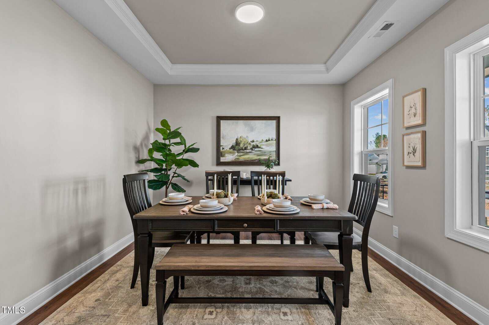 A dining room with a wooden table set for six, including plates, glasses, and napkins. Black chairs and a bench surround the table. A potted plant, wall art, and large windows brighten the neutral-toned space.