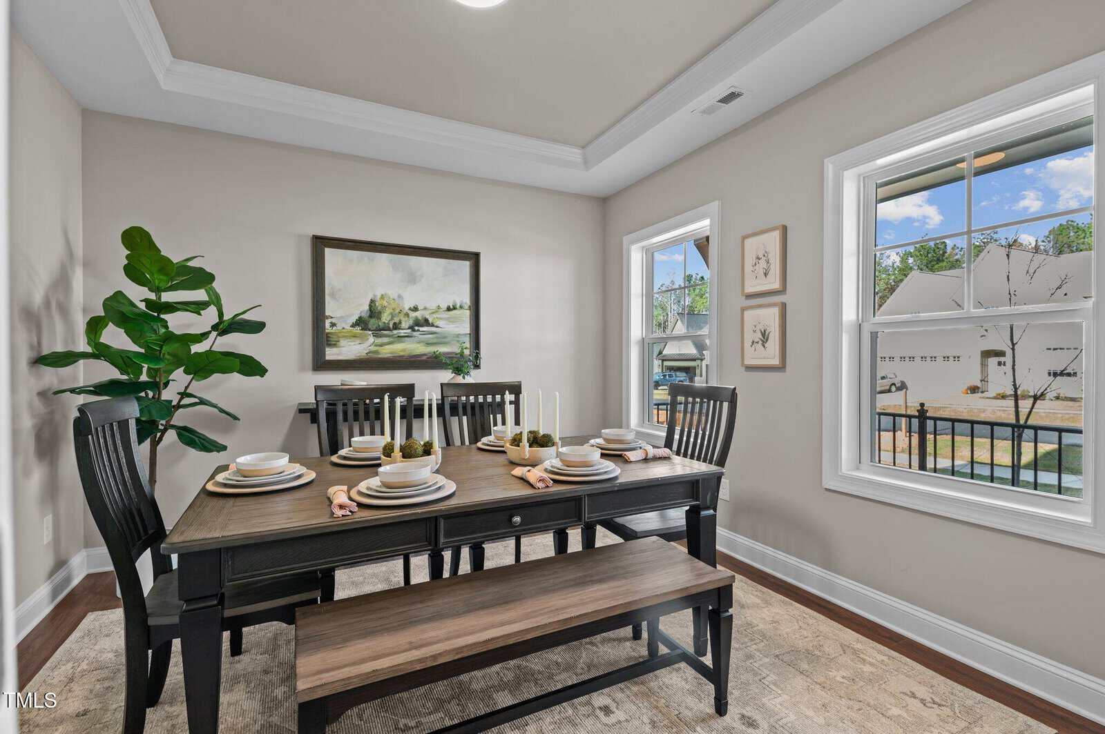 A bright dining room with a black table set for six, matching chairs, a wooden bench, a large plant, landscape artwork, and two windows showing an outdoor patio and pool.