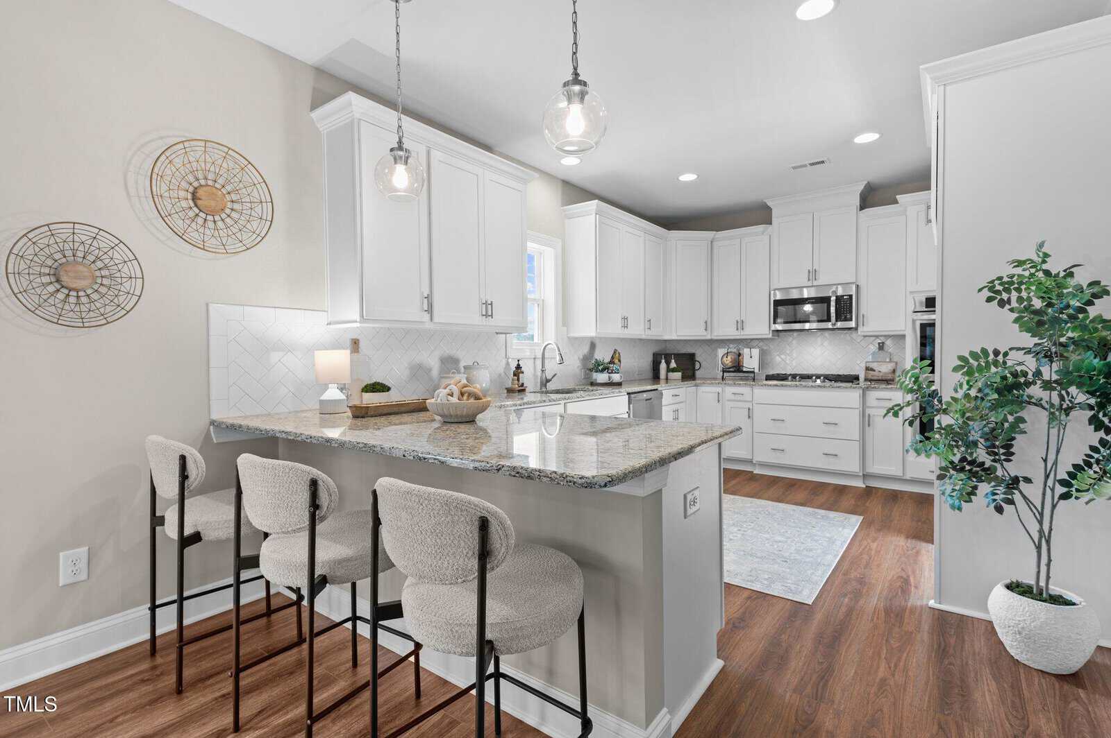 Modern kitchen with white cabinets, granite countertops, a kitchen island with three upholstered barstools, stainless steel appliances, wood flooring, pendant lights, wall decor, and a potted plant in the corner.