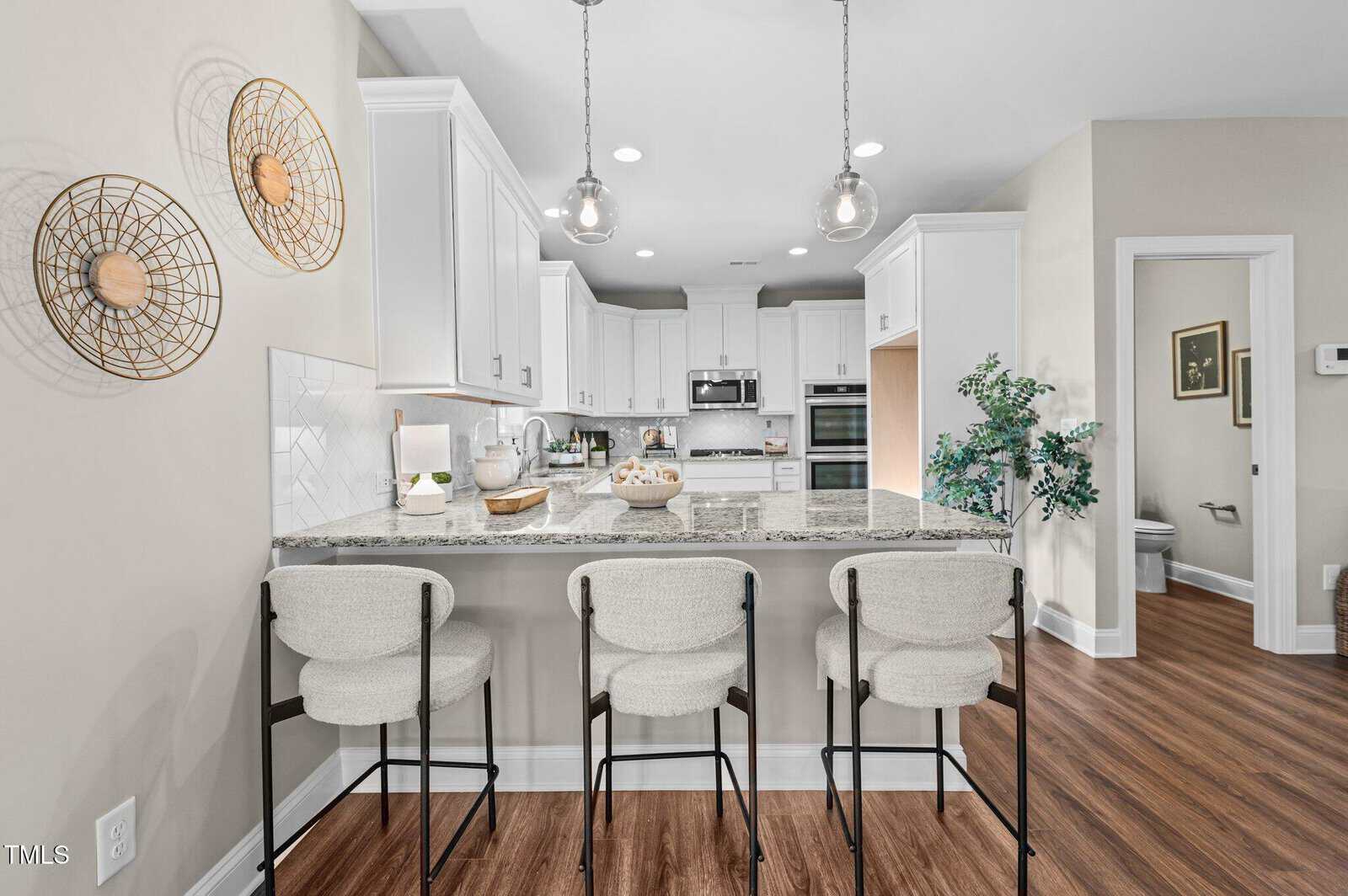 Modern kitchen with white cabinets, stainless steel appliances, granite island with three beige upholstered barstools, pendant lights above, and wood-look flooring. Decorative wall art and a partial view of a bathroom are visible.