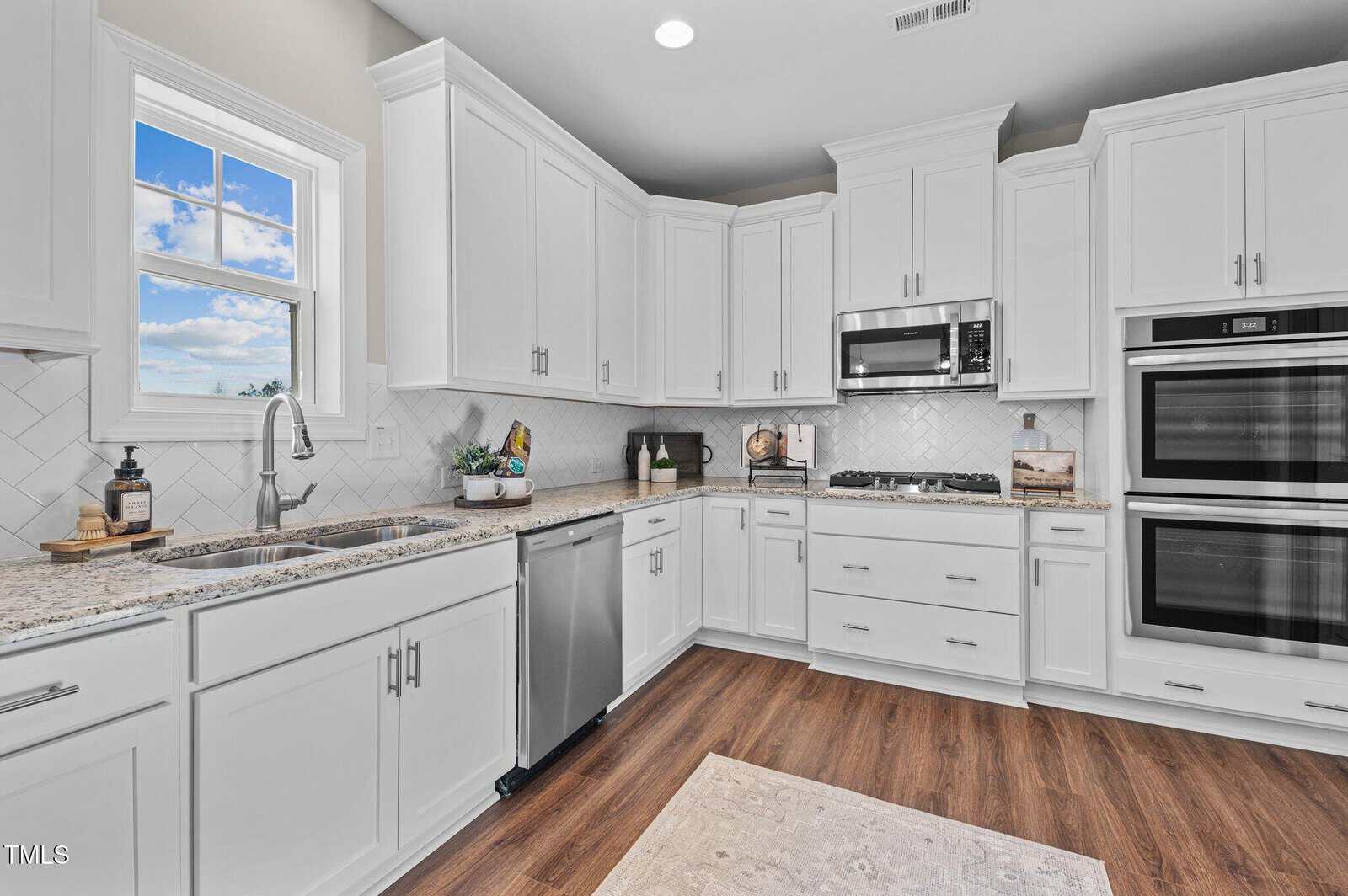 Bright, modern kitchen with white cabinets, stainless steel appliances, granite countertops, a window above the sink, hardwood floors, and a built-in microwave and double oven.
