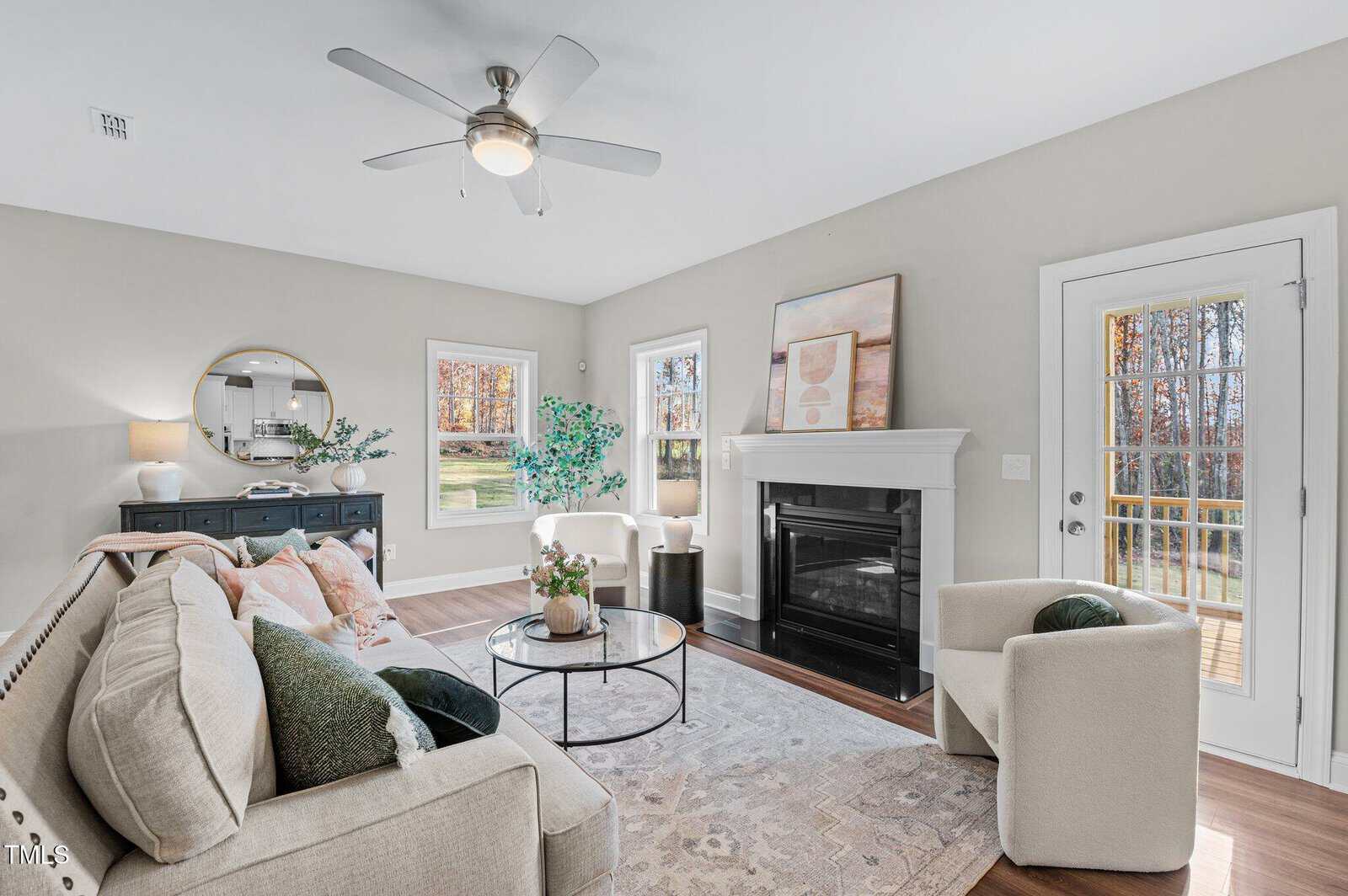 Bright living room with neutral furniture, a round coffee table, fireplace, artwork, large windows, and a glass door. A round mirror and greenery add decorative touches. Light wood floors and soft rug complete the cozy space.