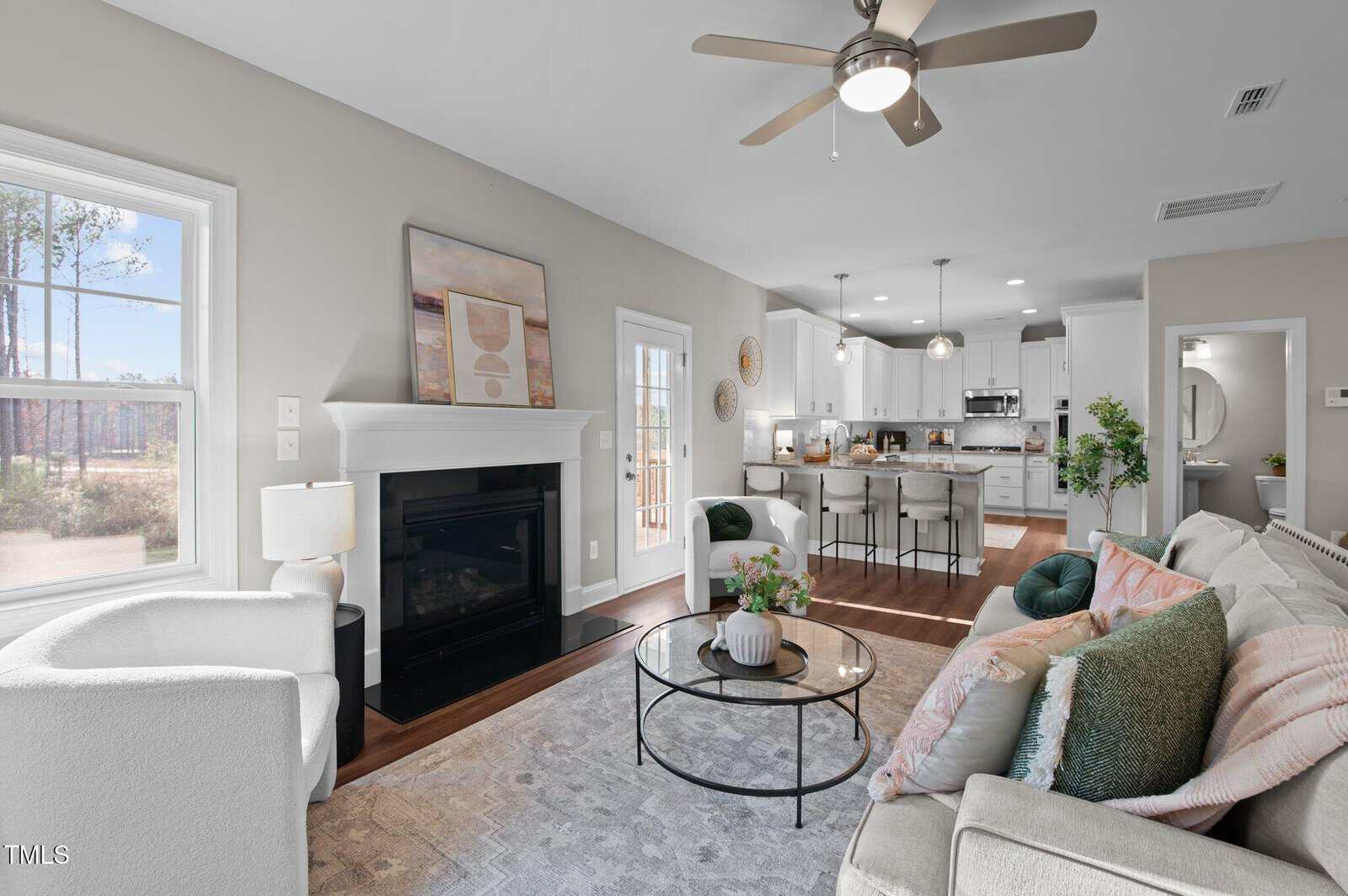Modern living room with white armchairs, gray sofa, round coffee table, and a fireplace. Open layout leads to a bright kitchen with an island and barstools. Ceiling fans and large windows add light.