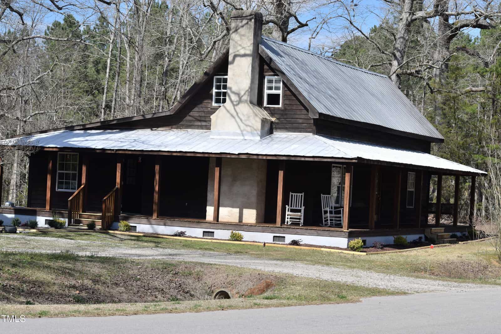 A rustic wooden cabin with a metal roof and a wraparound porch featuring two rocking chairs. The cabin is surrounded by bare trees and is located near a paved road. The sky is clear and sunny.