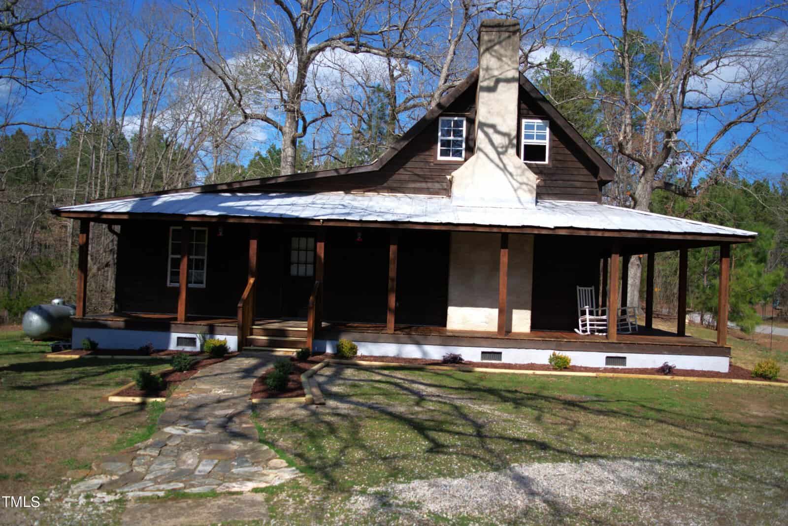 A rustic wooden cabin with a metal roof and a wraparound porch featuring a rocking chair. Surrounding the cabin are sparse trees and a grassy lawn. A stone path leads to the porch steps, and the sky is clear with a few clouds.