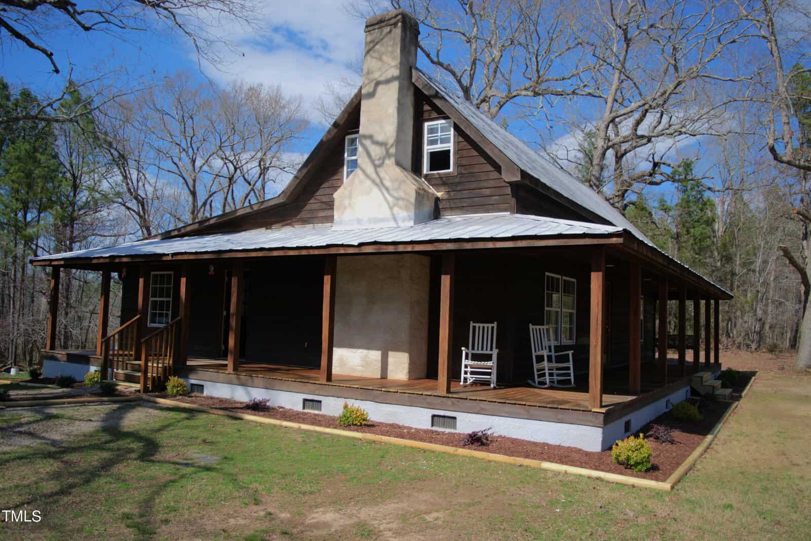 A rustic, wooden cabin with a wraparound porch and a metal roof, surrounded by leafless trees. Two white rocking chairs sit on the porch, and the sky is blue with scattered clouds.
