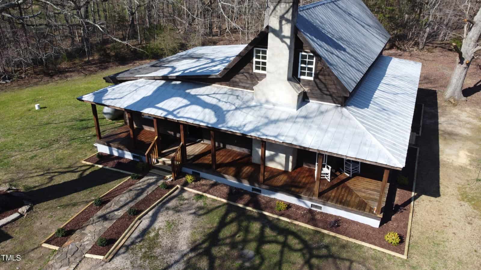 Aerial view of a rustic wooden house at 2603 Moss Ledford Road, Franklinton, featuring a metal roof and wraparound porch. Surrounded by bare trees and a grassy yard, a stone pathway leads to the entrance while shadows of branches stretch across the roof and lawn.