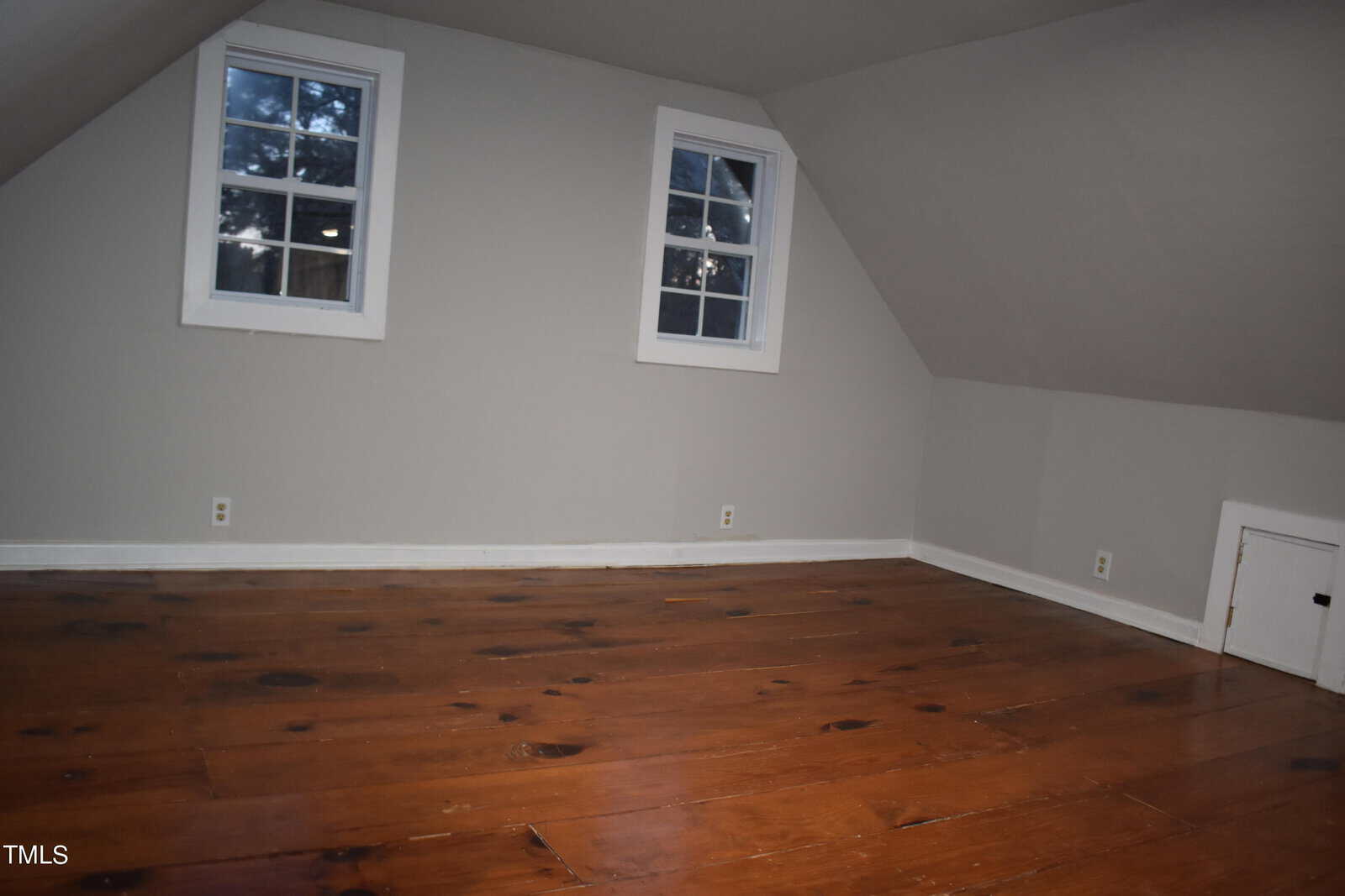 An empty attic room with sloped ceilings and two small windows. The walls are painted light gray, and the floor is made of dark polished wood. A small square door is visible on the right wall.