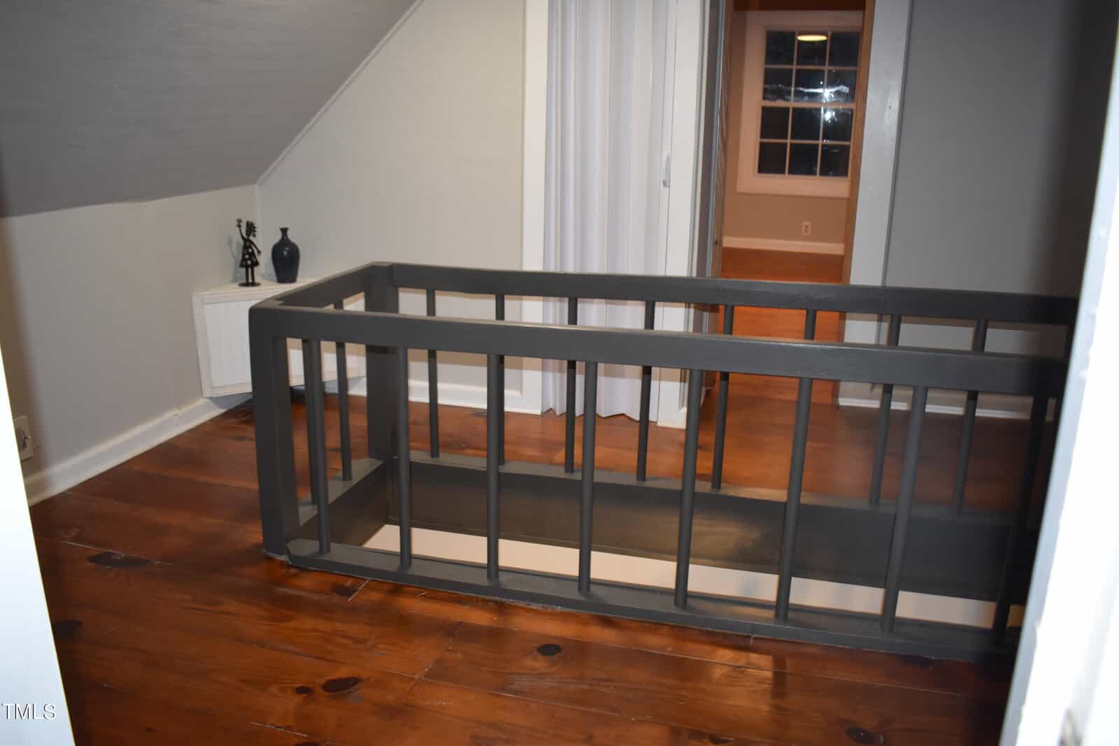 A wooden staircase railing in a gray-painted room with hardwood floors. Two small decorative items are on the left near a wall. A door in the background is slightly ajar, revealing another room with a window.
