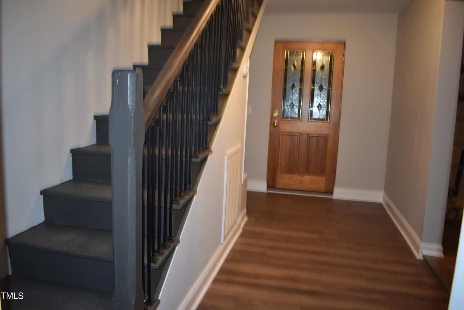 A hallway with wooden flooring features a staircase on the left with dark railings and steps. To the right, a wooden door with decorative glass panels is visible. Walls are painted light gray, contributing to a modern, inviting atmosphere.