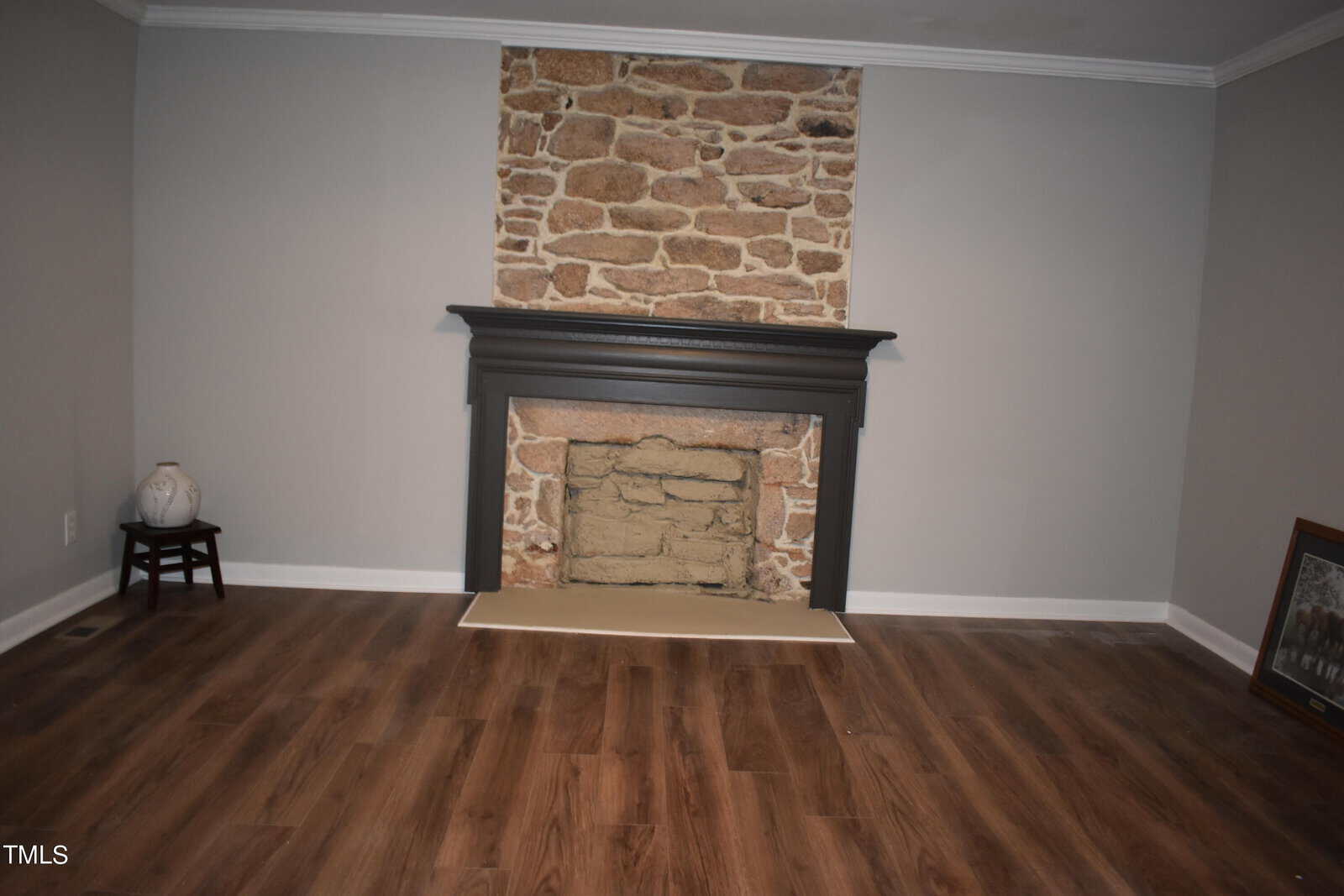 An empty room with a stone fireplace and a dark wooden mantel. The walls are painted gray, and the floor has a wood-like finish. A small stool with a vase is on the left, and a framed picture leans against the wall on the right.