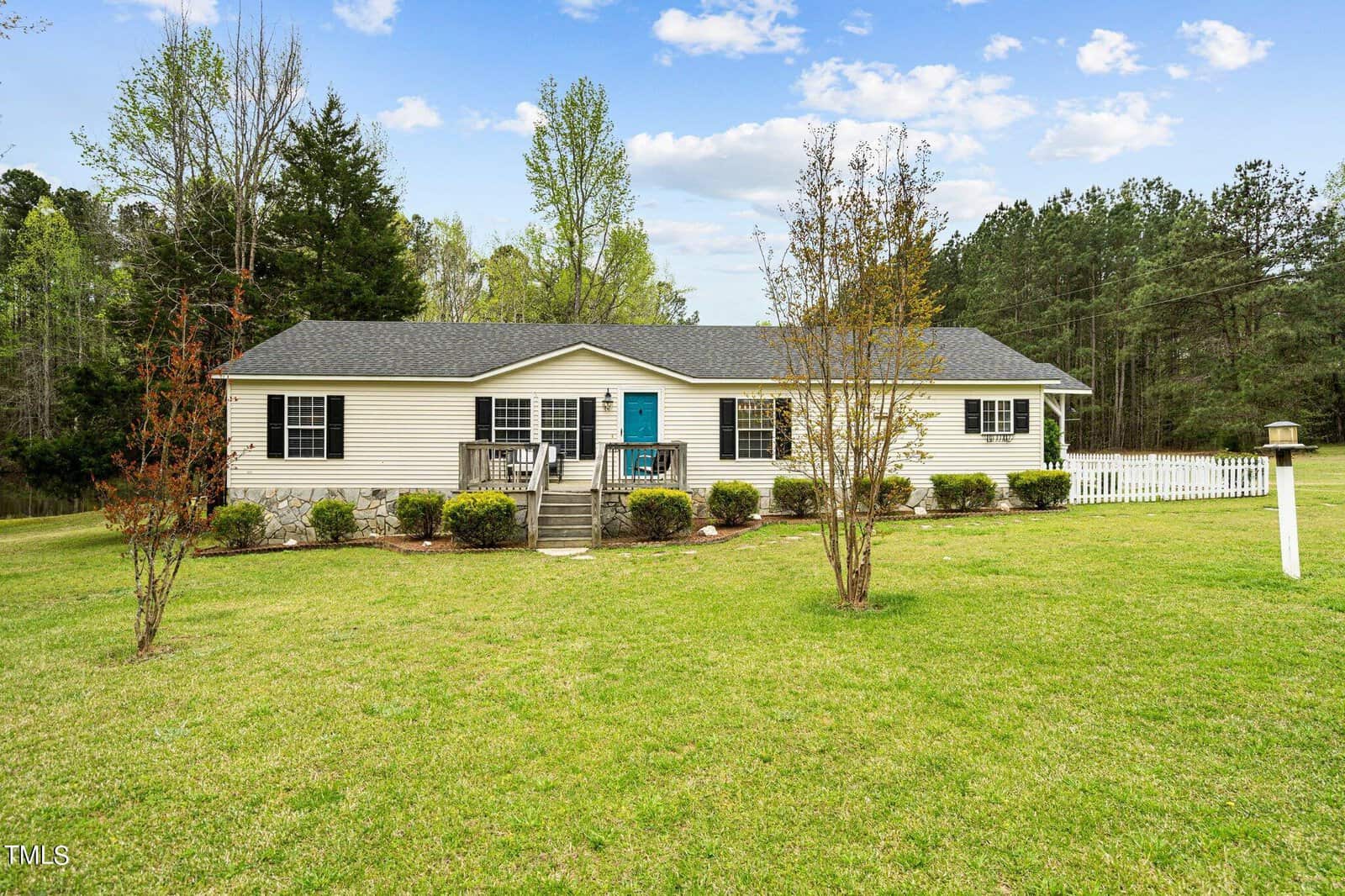 A single-story house at 3918 Batson Drive in Franklinton features a blue door, surrounded by lush green grass and trees. A small stairway leads to the entrance, with a white fence on the right. The sky above is partly cloudy.