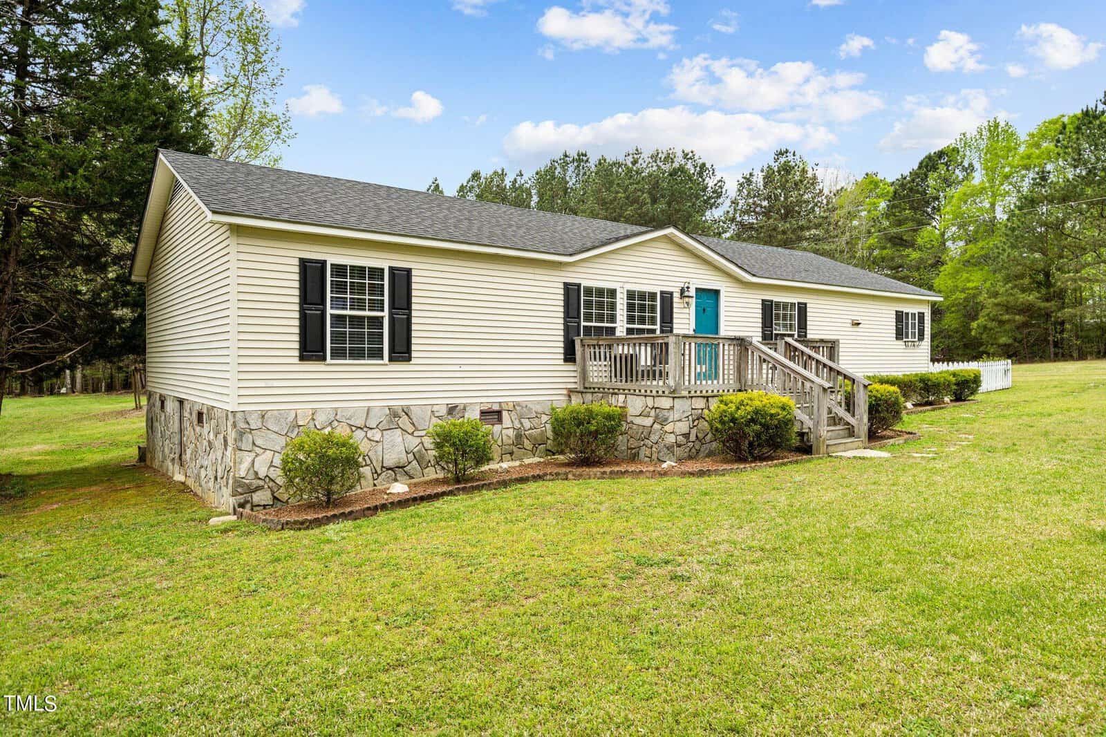 A single-story house with beige siding and a stone foundation, featuring a teal front door and black shutters. A wooden porch with steps leads to the entrance. It's surrounded by a grassy lawn and trees in the background under a partly cloudy sky.