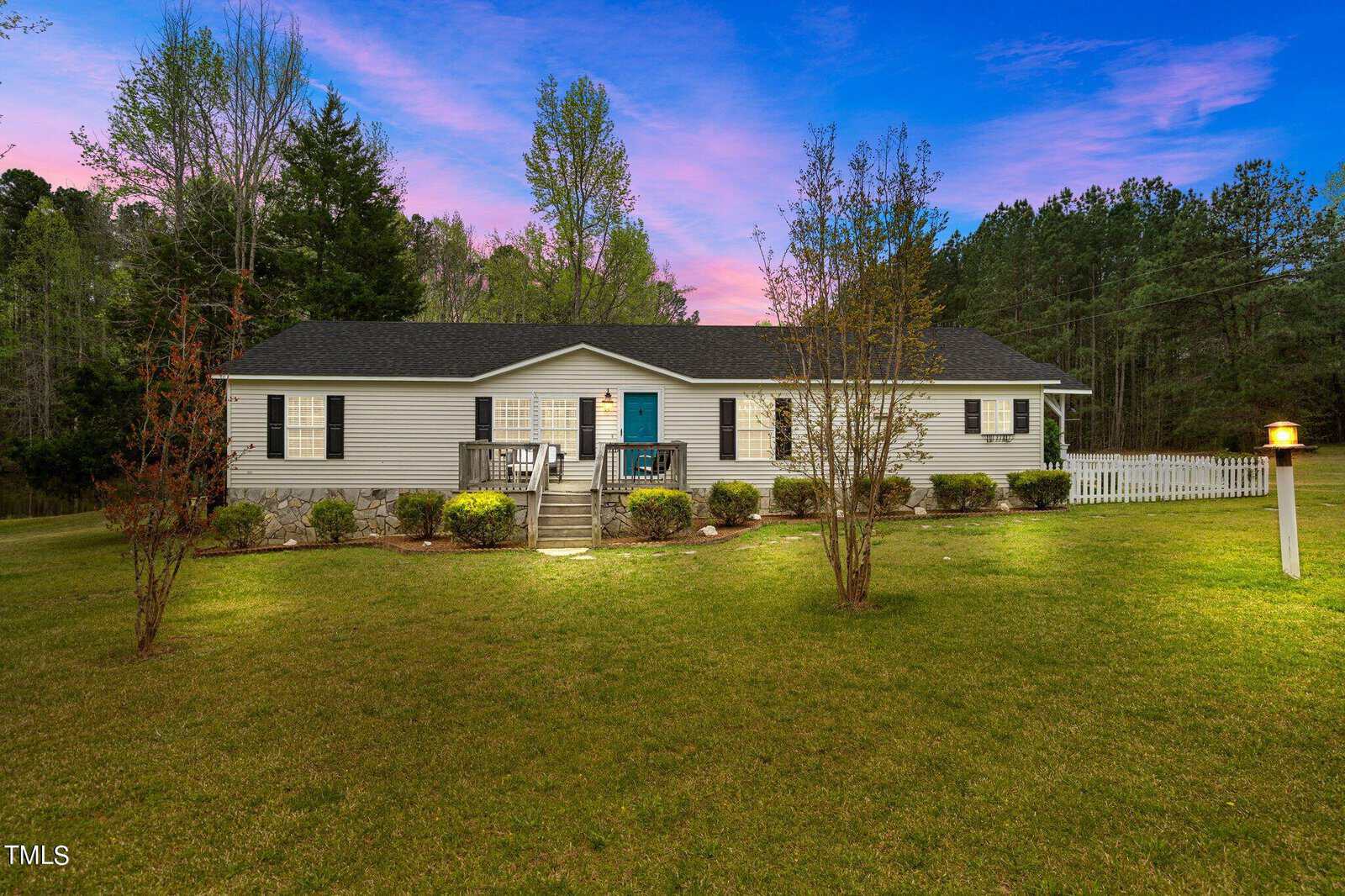 A single-story house with a blue door is surrounded by a manicured lawn and path lights. Trees and shrubs frame the house. The sky is a blend of pink, purple, and blue hues, suggesting sunset. A white fence is visible on the right.