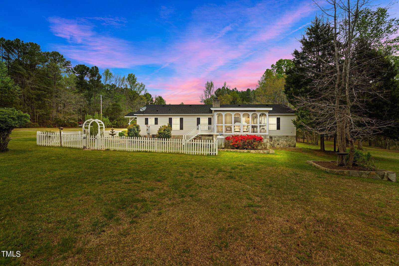 A quaint white house with a large porch and surrounding green lawn. In the foreground, there's a white picket fence and garden archway, with sunset skies painted in vibrant pink and blue hues overhead.