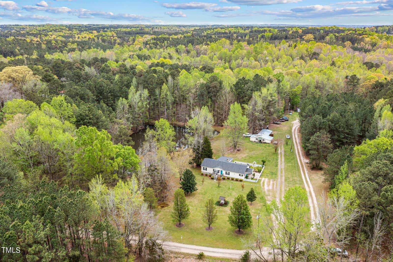 Aerial view of a rural property surrounded by dense forest. A white house with a long driveway is centered in the image. Several vehicles are parked along the driveway, and a small pond is visible to the left. The sky is partly cloudy.