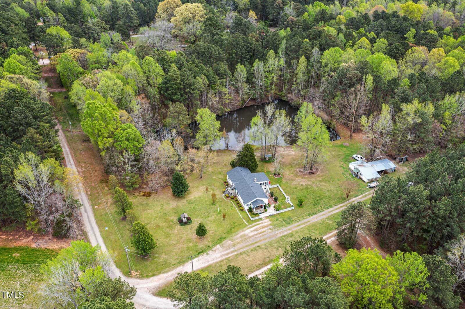 Aerial view of a rural landscape featuring a house with a gray roof surrounded by trees. A dirt road leads to the property, which includes a pond and a small outbuilding. Forested areas and patches of grass are visible throughout the scene.
