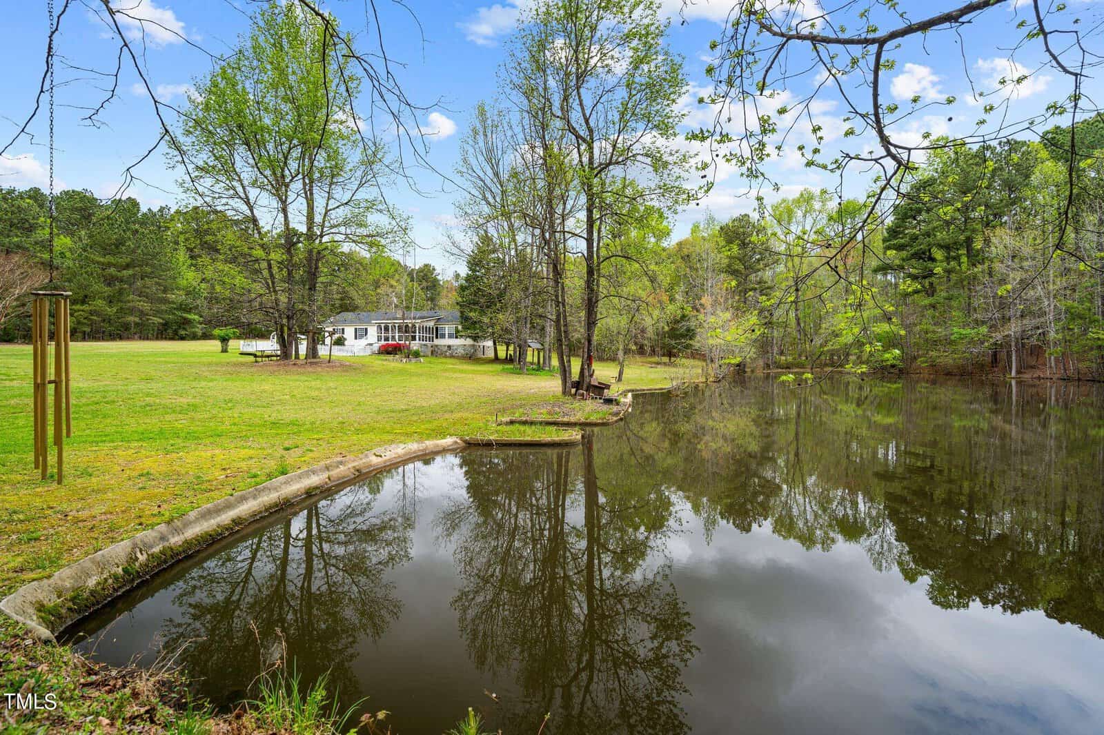 A tranquil lakeside scene with a reflective pond, surrounded by lush green grass and trees. In the background, there's a white house with a porch. The sky is clear with a few clouds.