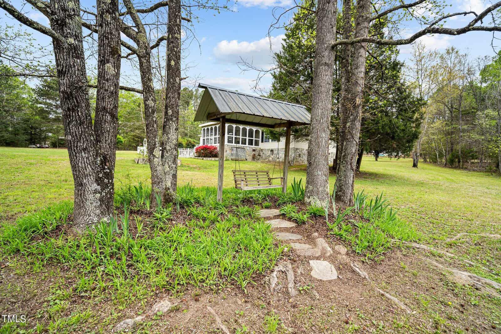 A wooden swing with a green metal roof is nestled among trees in a grassy area. A stone path leads to the swing, with lush foliage surrounding it. In the background, there's a white house with large windows and blooming red bushes.
