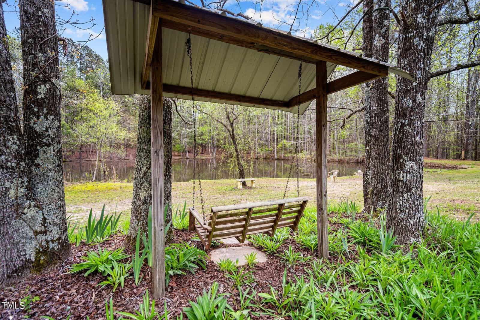 A wooden swing hangs under a small roofed structure surrounded by tall trees and lush greenery. In the background, a serene pond and a stone table add to the tranquil outdoor setting.